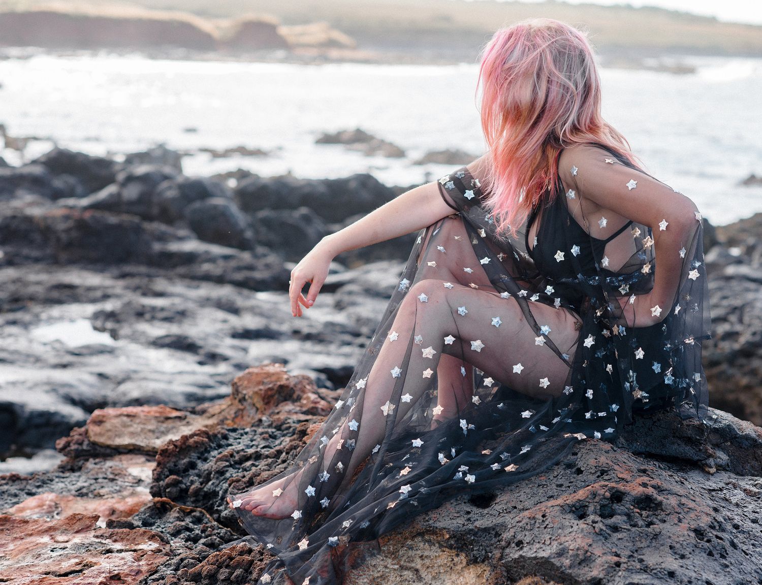 Person in flowing polka dot dress sitting on rocky beach at sunset.