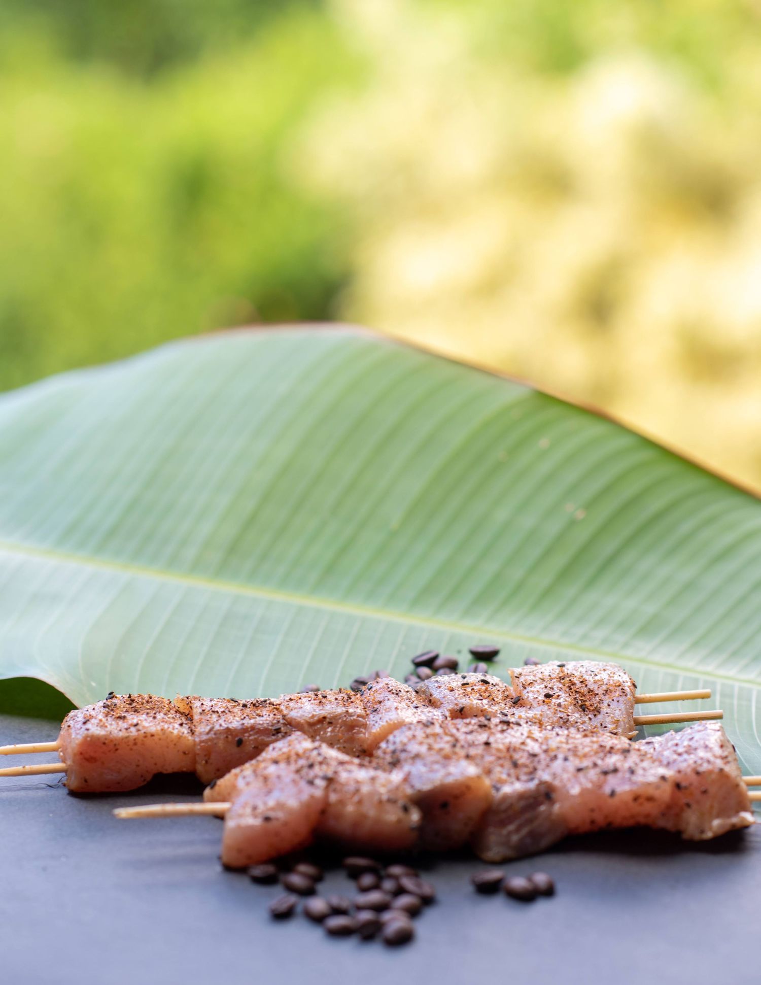 Raw pork tenderloin skewers with seasoning resting on a banana leaf with coffee beans scattered around.