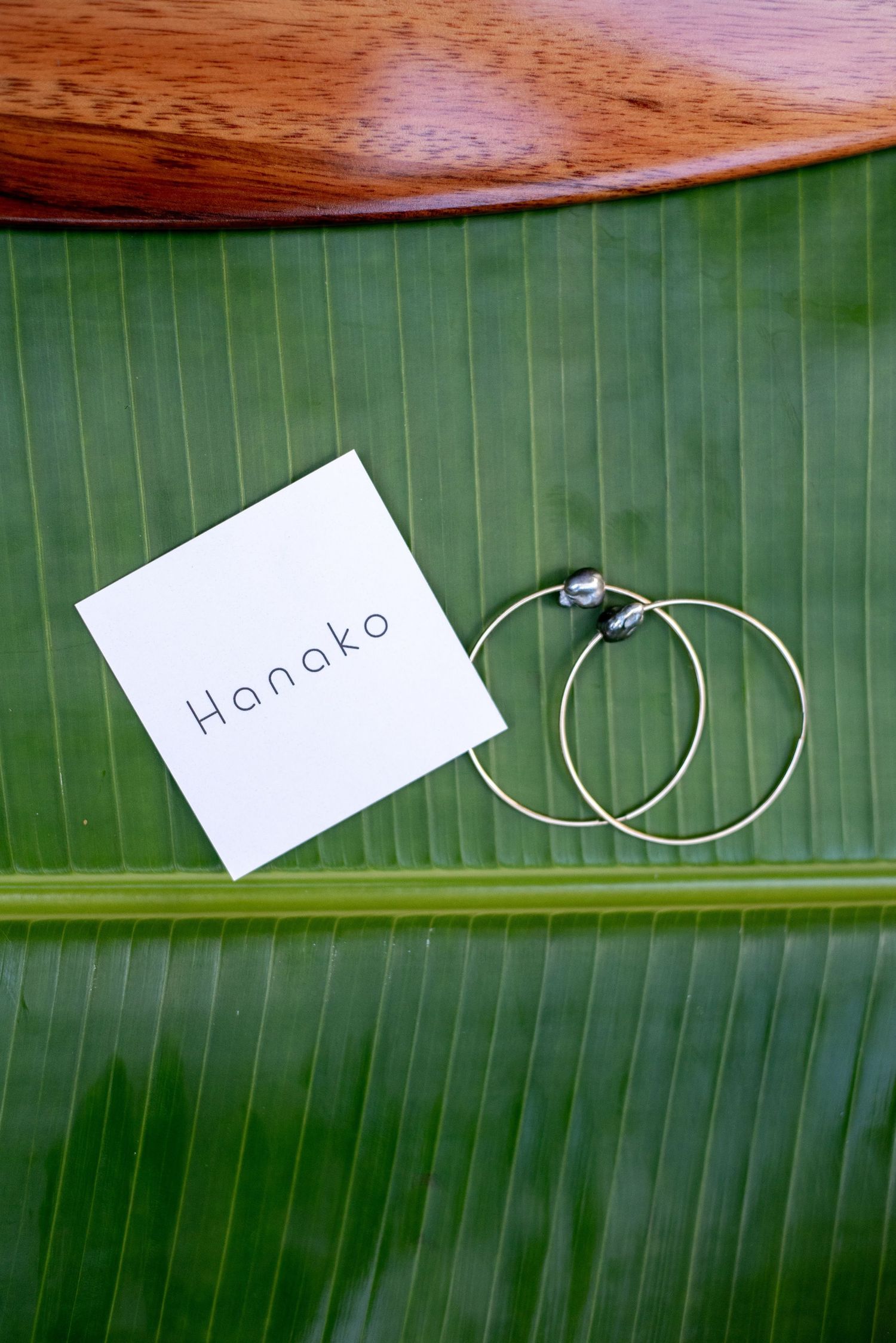 Silver hoop earrings and a Hanako branded card displayed on a large green banana leaf surface.