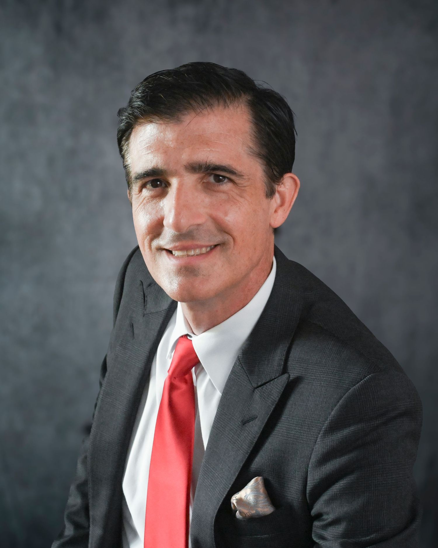 Professional portrait of a man in a grey suit with a bright red tie and white pocket square against a dark background.