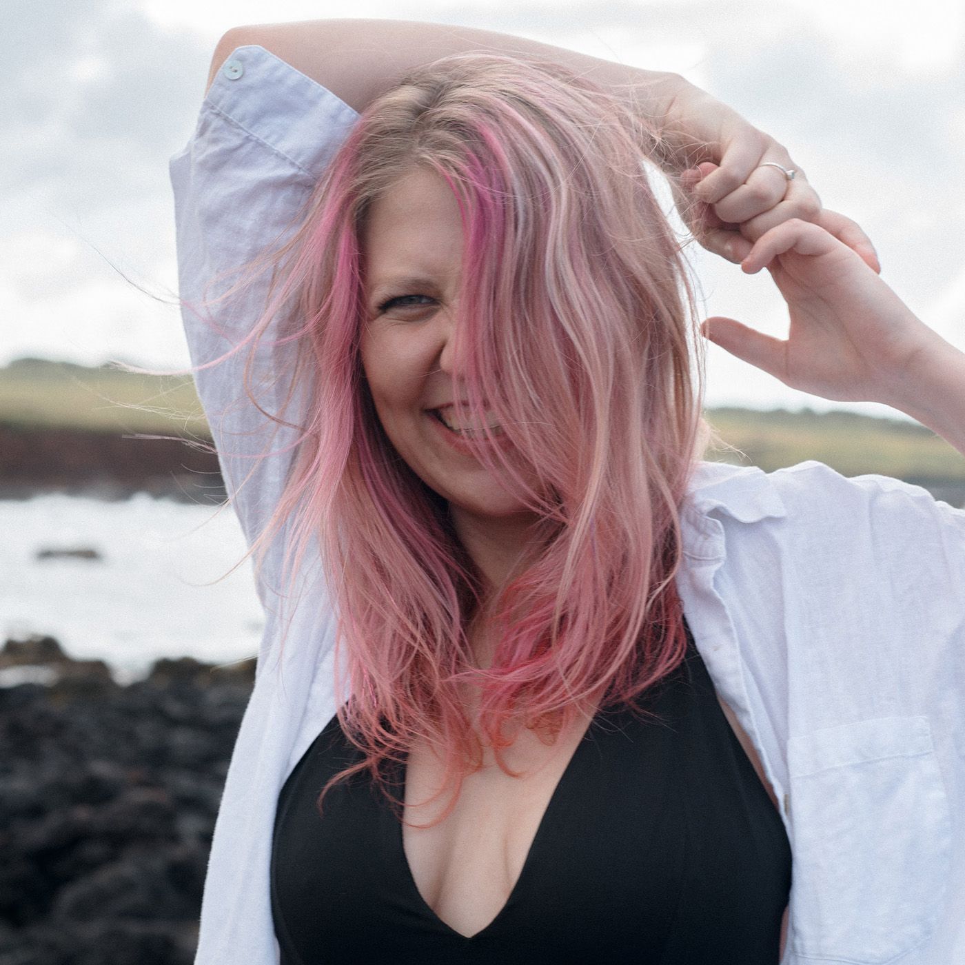 Someone with pink hair smiling and posing outdoors near a rocky coastline.