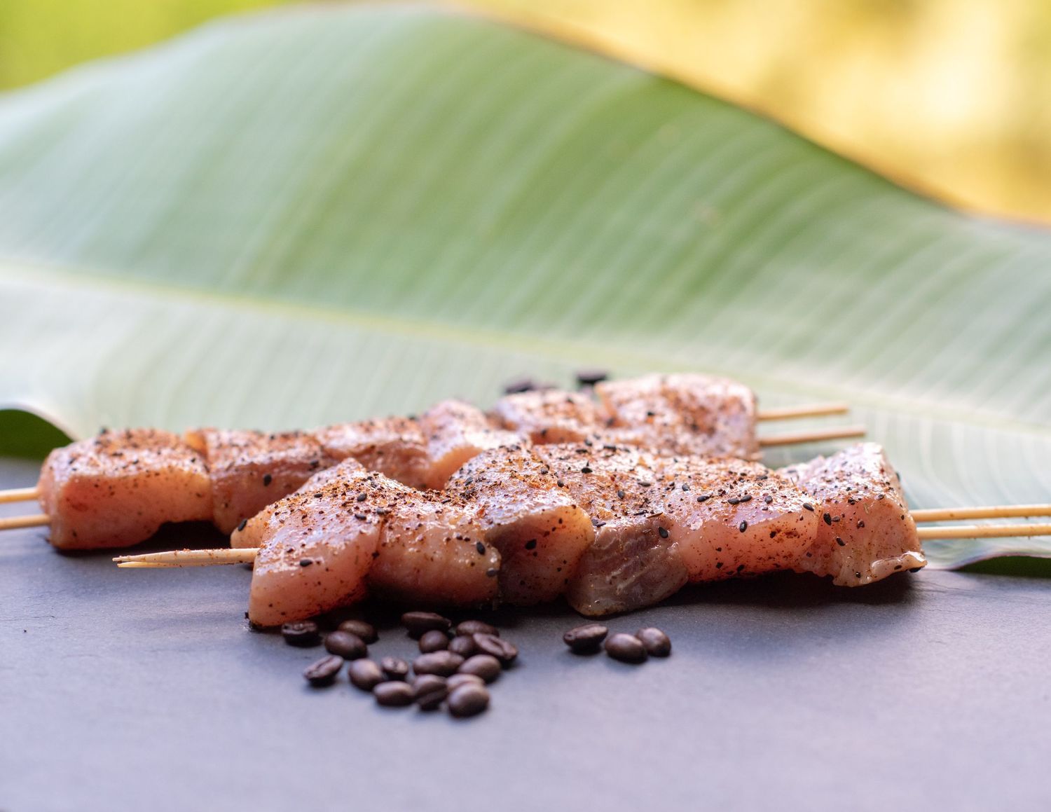 Raw skewered chicken pieces with peppercorns and spices on a banana leaf with blurred yellow background.