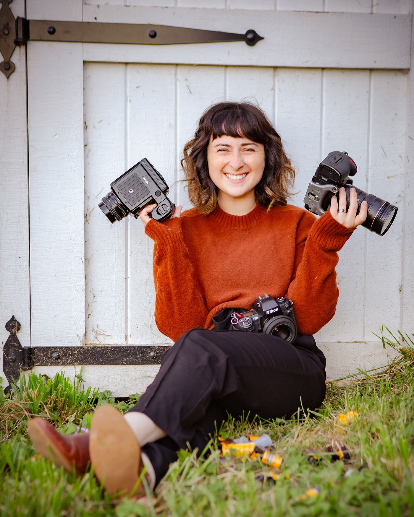 Photographer holding multiple cameras while sitting against white barn door.