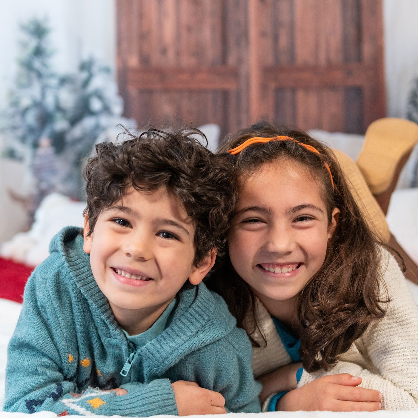 Two children in winter sweaters smile while lying on holiday-themed blankets.