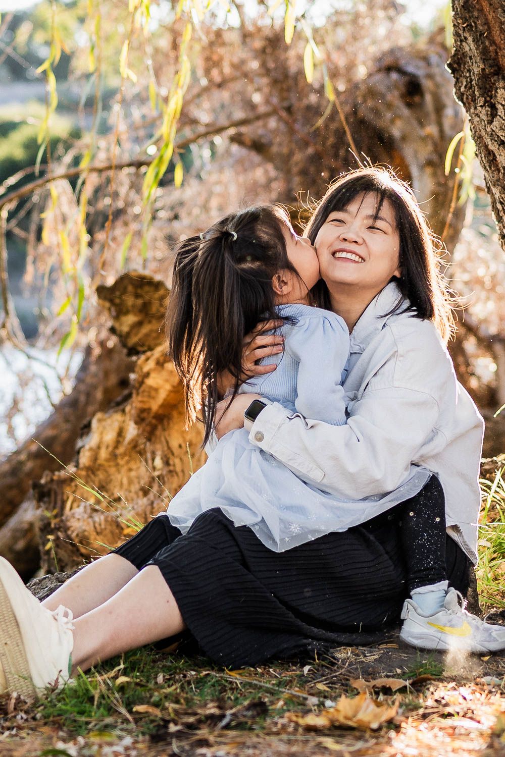 A mother and child share a joyful, intimate moment while sitting on grass under spring willow tree.