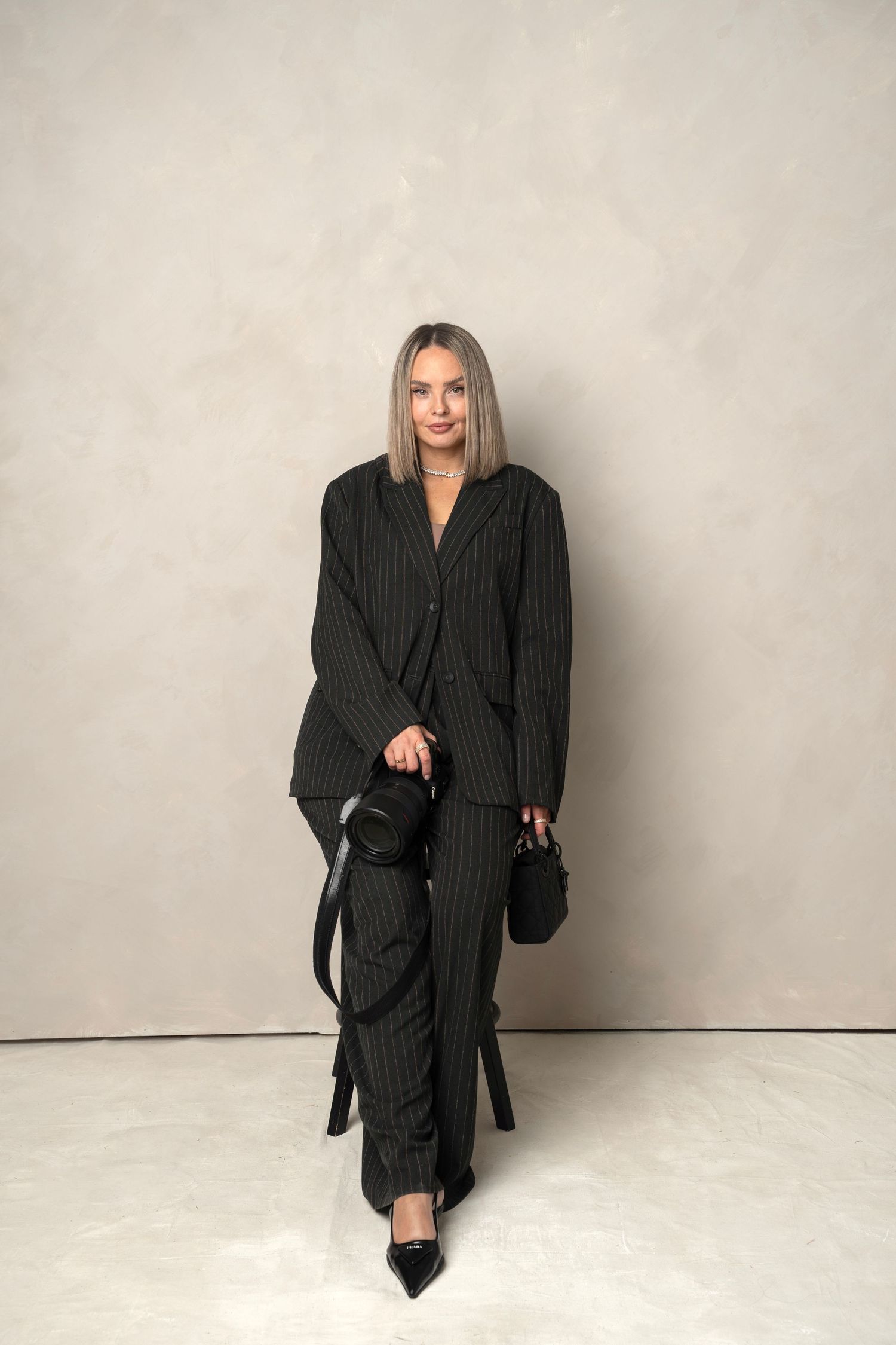 A person wearing an oversized black pinstripe suit ensemble with pointed heels and a small handbag against a neutral backdrop.