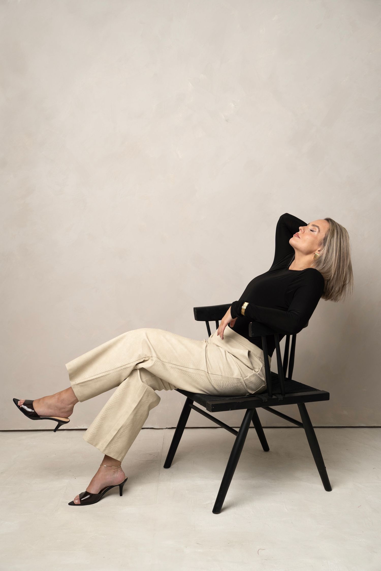 Person in black top and beige pants reclines on a black wooden chair against neutral backdrop.