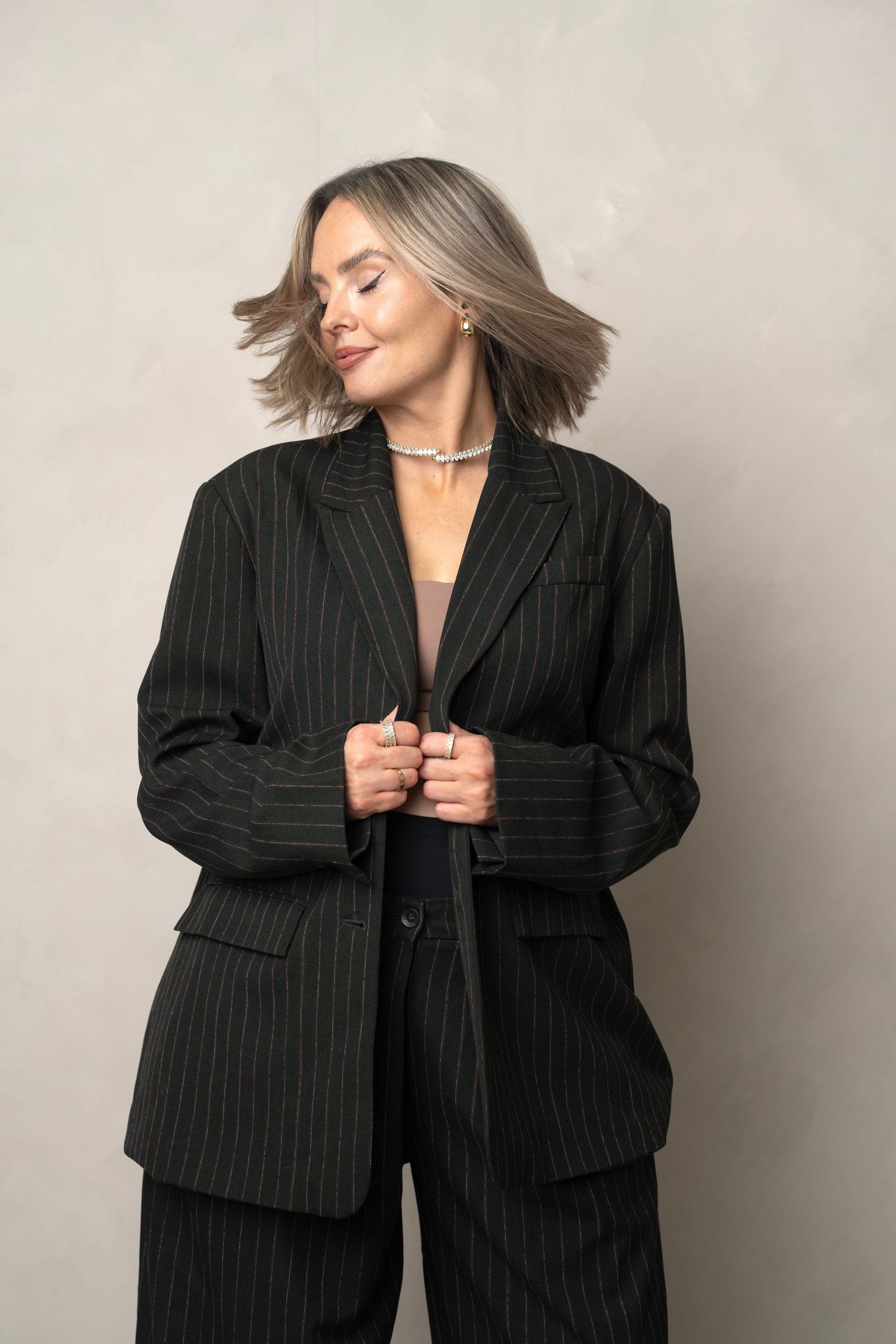 Fashion portrait of person in black pinstripe power suit with ash blonde hair blowing in wind against neutral background.