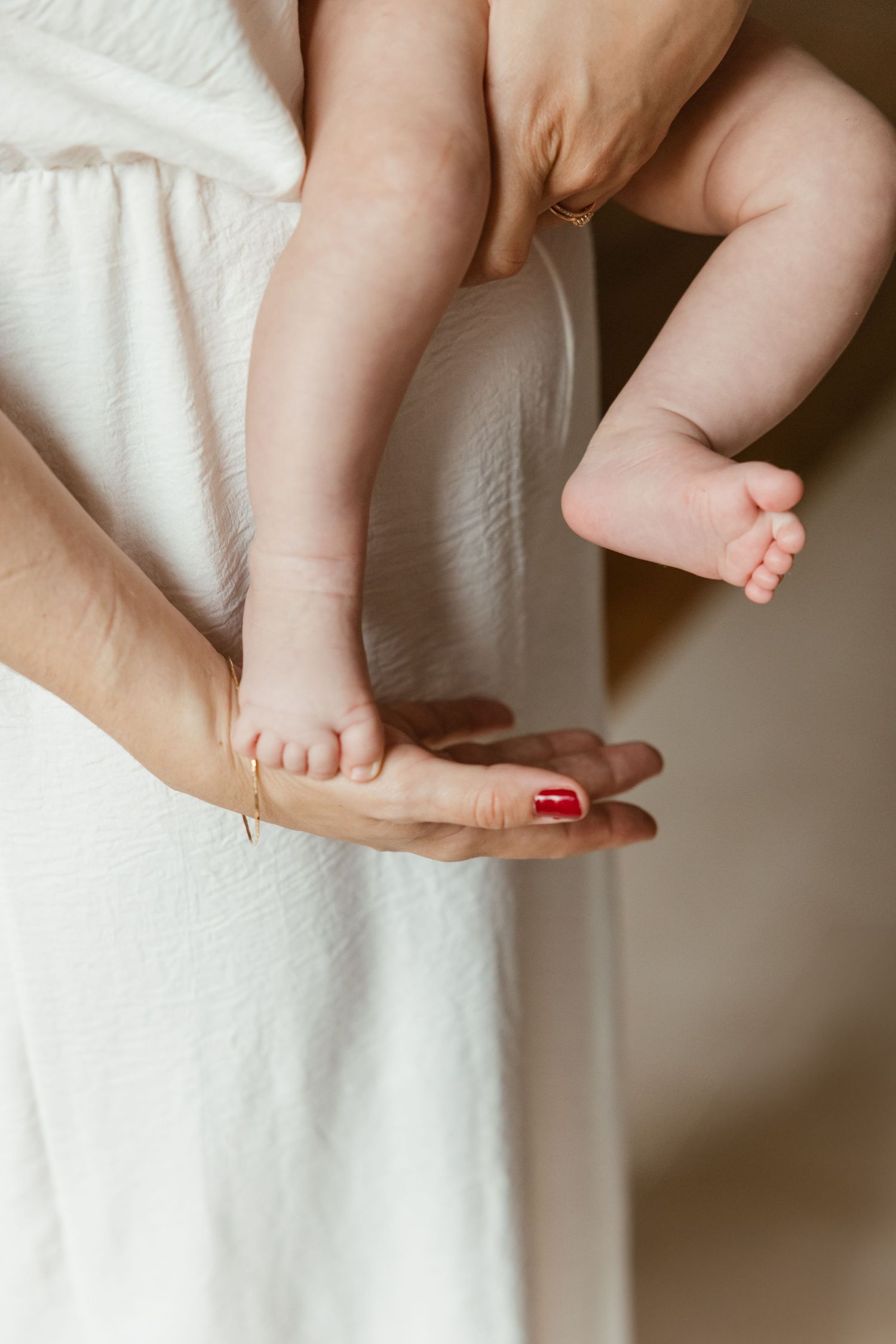 Close-up view of tiny baby feet resting on adult hands with red manicured nails against white fabric background.