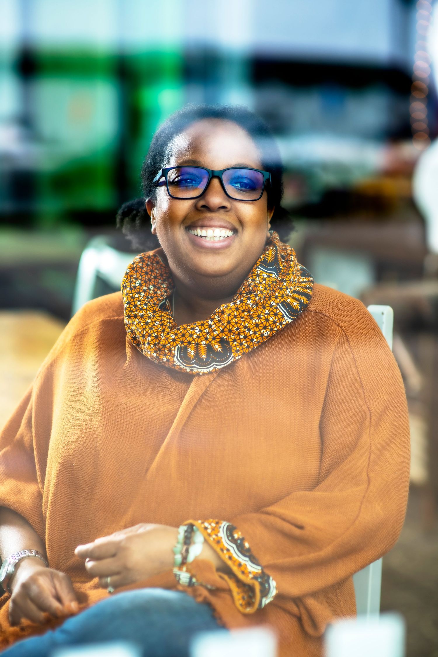 A person in a tan sweater smiles while wearing blue glasses and an ornate gold statement necklace.