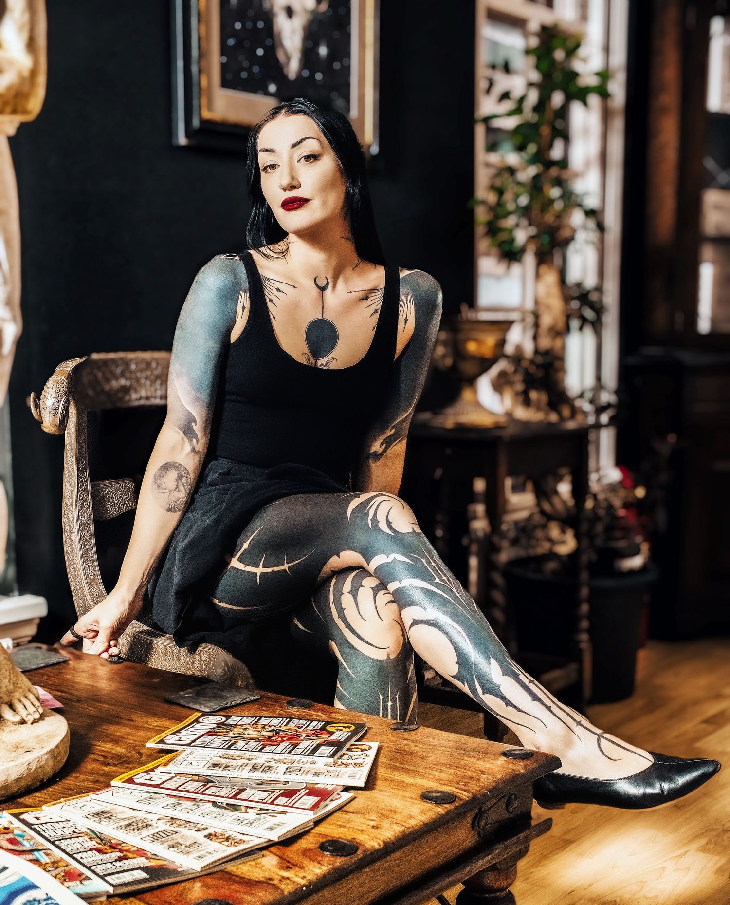 A tattooed woman in black athletic wear sits on a wooden chair with vintage posters on a table.