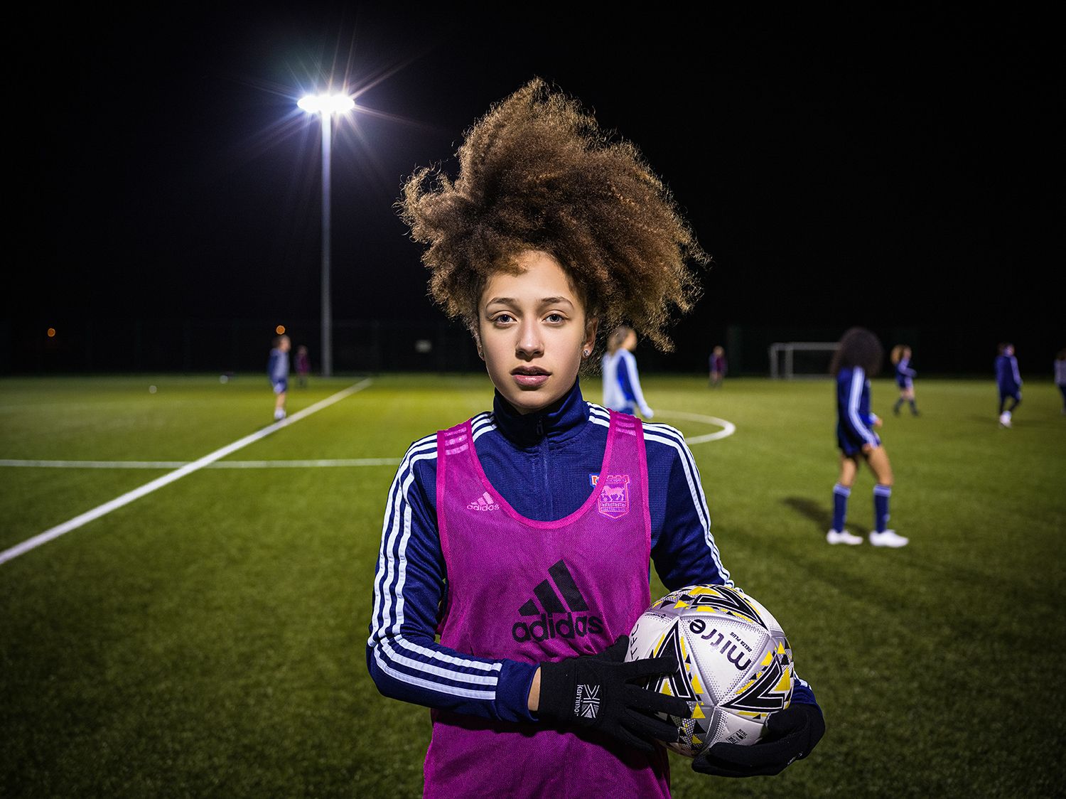 A soccer player in a purple training bib holds a ball under stadium lights during nighttime practice.