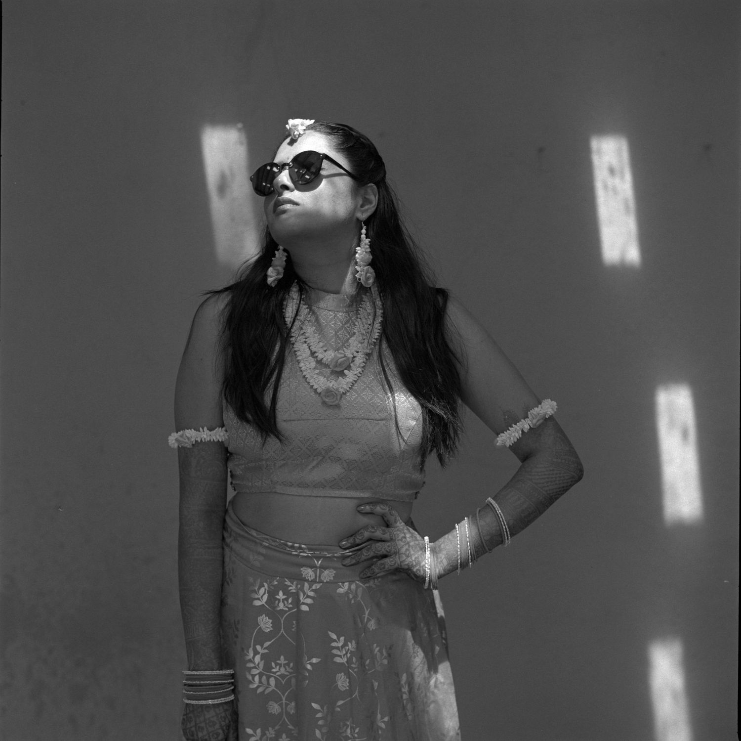 Black and white portrait of someone in sunglasses and jewelry posed against a light wall with shadows.