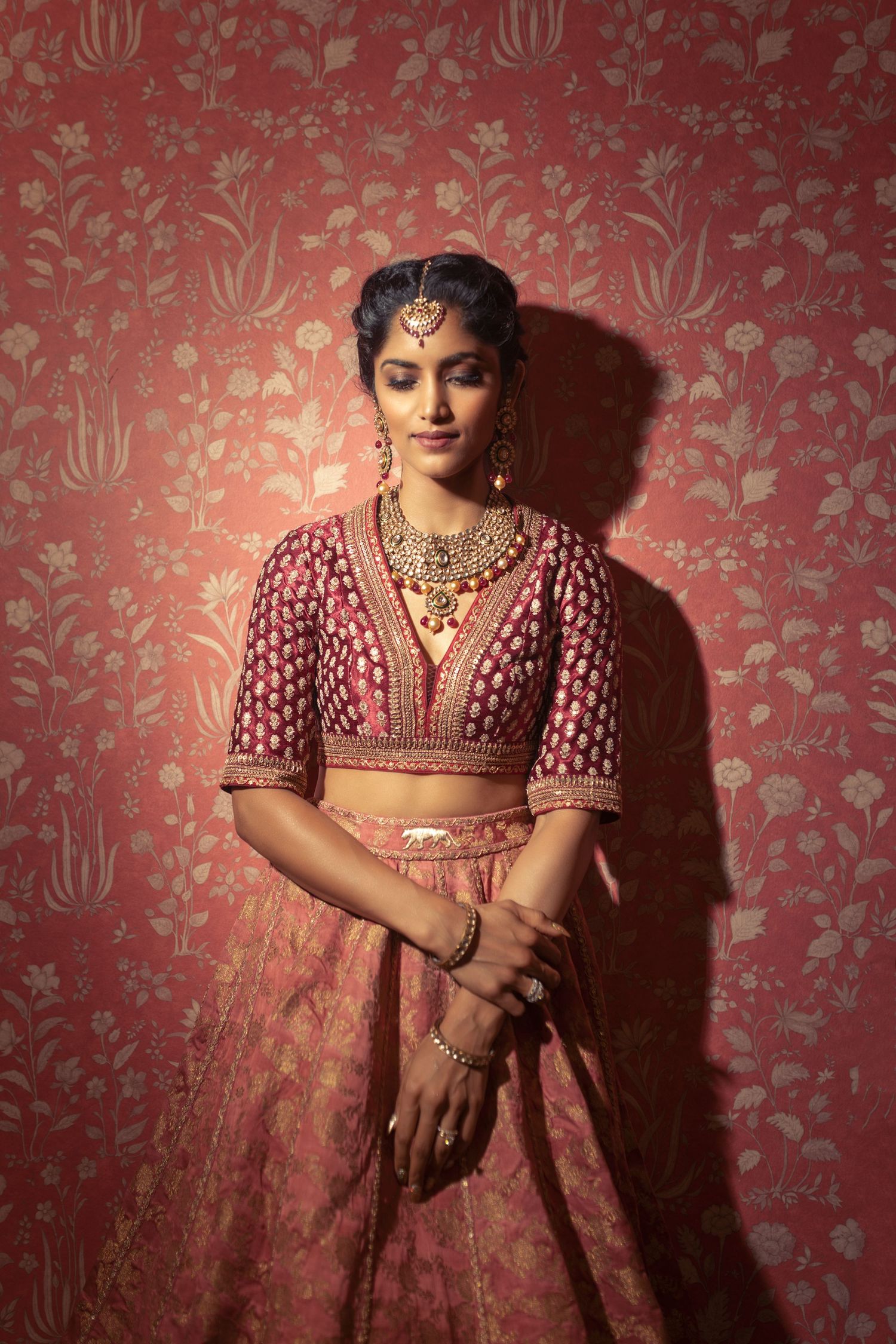 Traditional Indian bridal attire featuring a pink embellished lehenga and gold jewelry against a floral red backdrop.