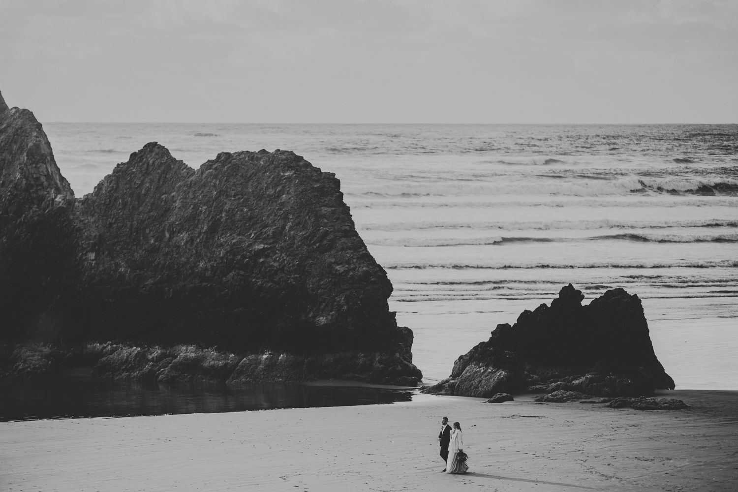 A black and white coastal landscape with silhouetted figures standing between dramatic rock formations on a sandy beach.