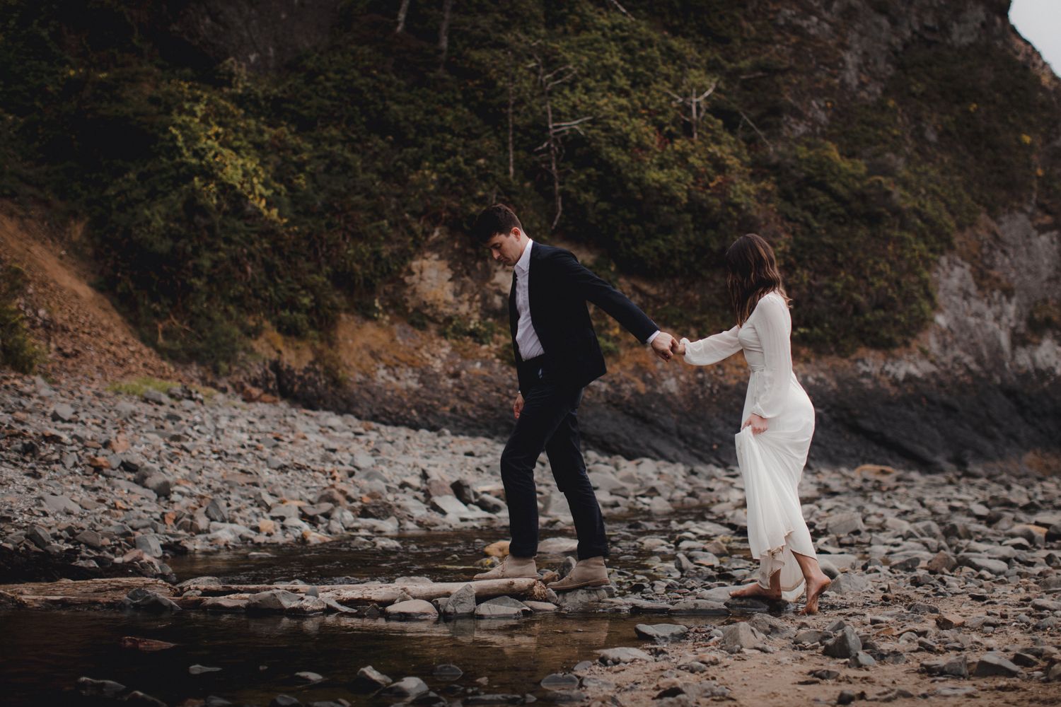Two people in formal attire hold hands while carefully crossing rocky coastal terrain at dusk.