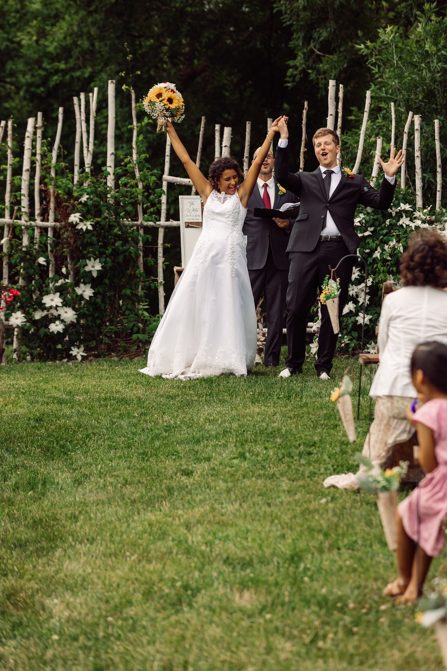 Joyful newlyweds celebrating with raised arms in front of a rustic wedding backdrop.