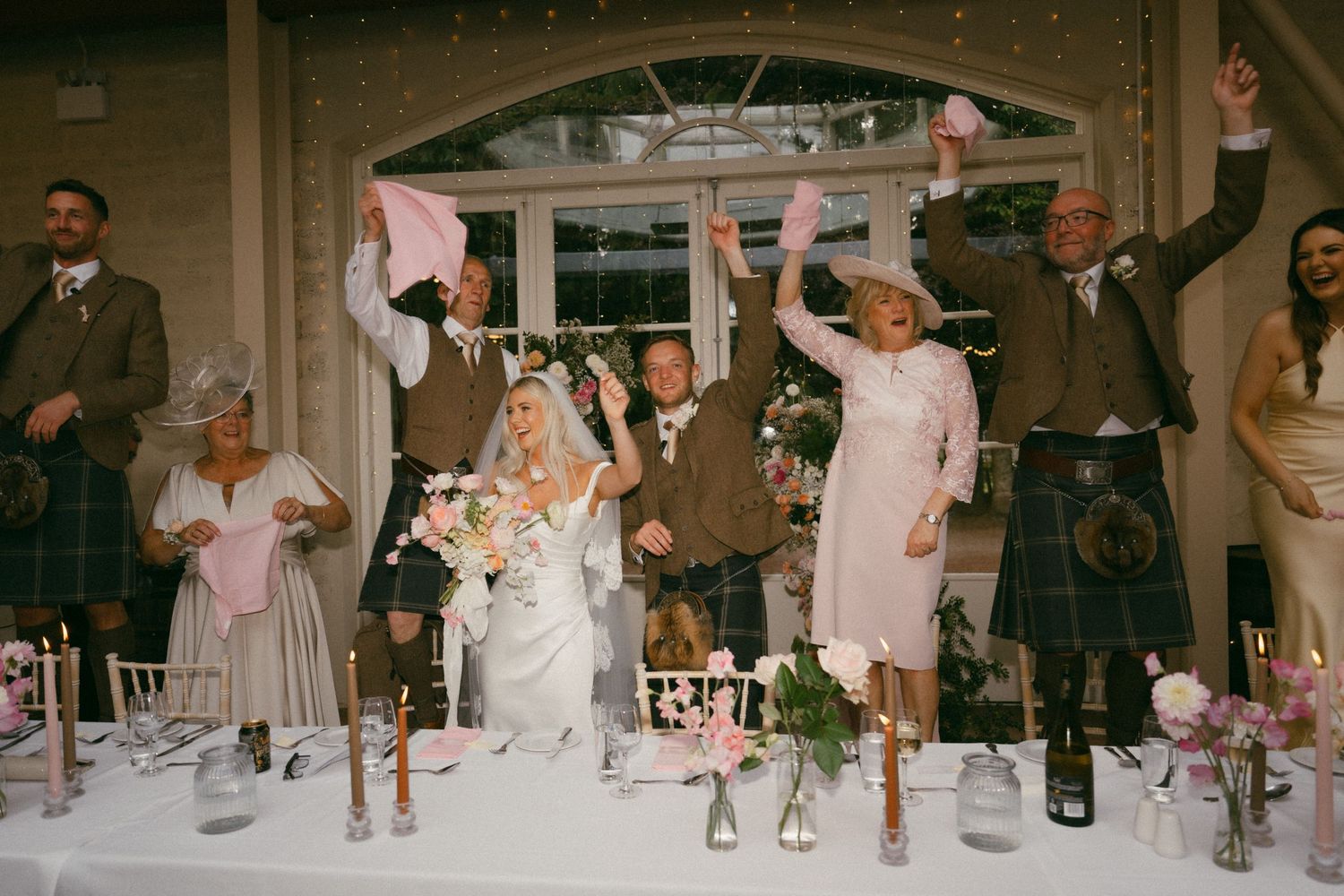 Wedding reception guests celebrate and cheer enthusiastically at the head table during a joyous dinner celebration.