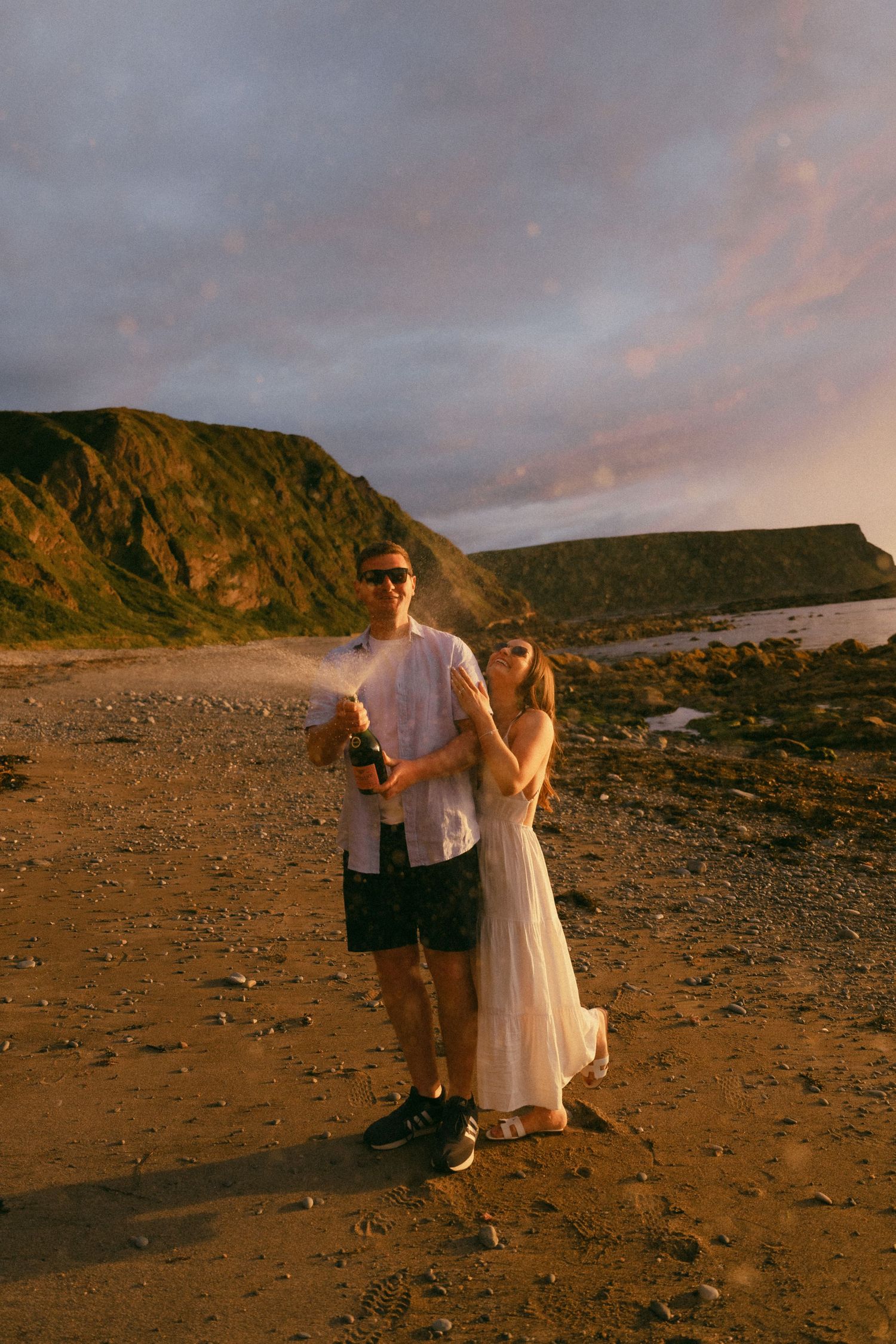 A couple stands on a rocky beach at sunset with dramatic coastal cliffs and golden light in the background.