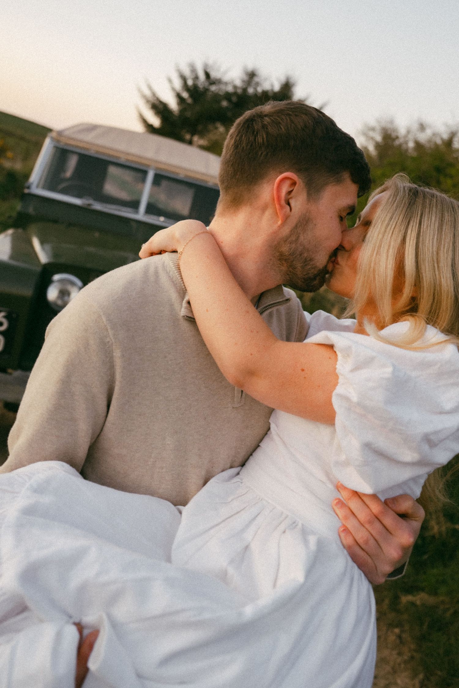 A romantic moment captured in front of a vintage car during sunset.