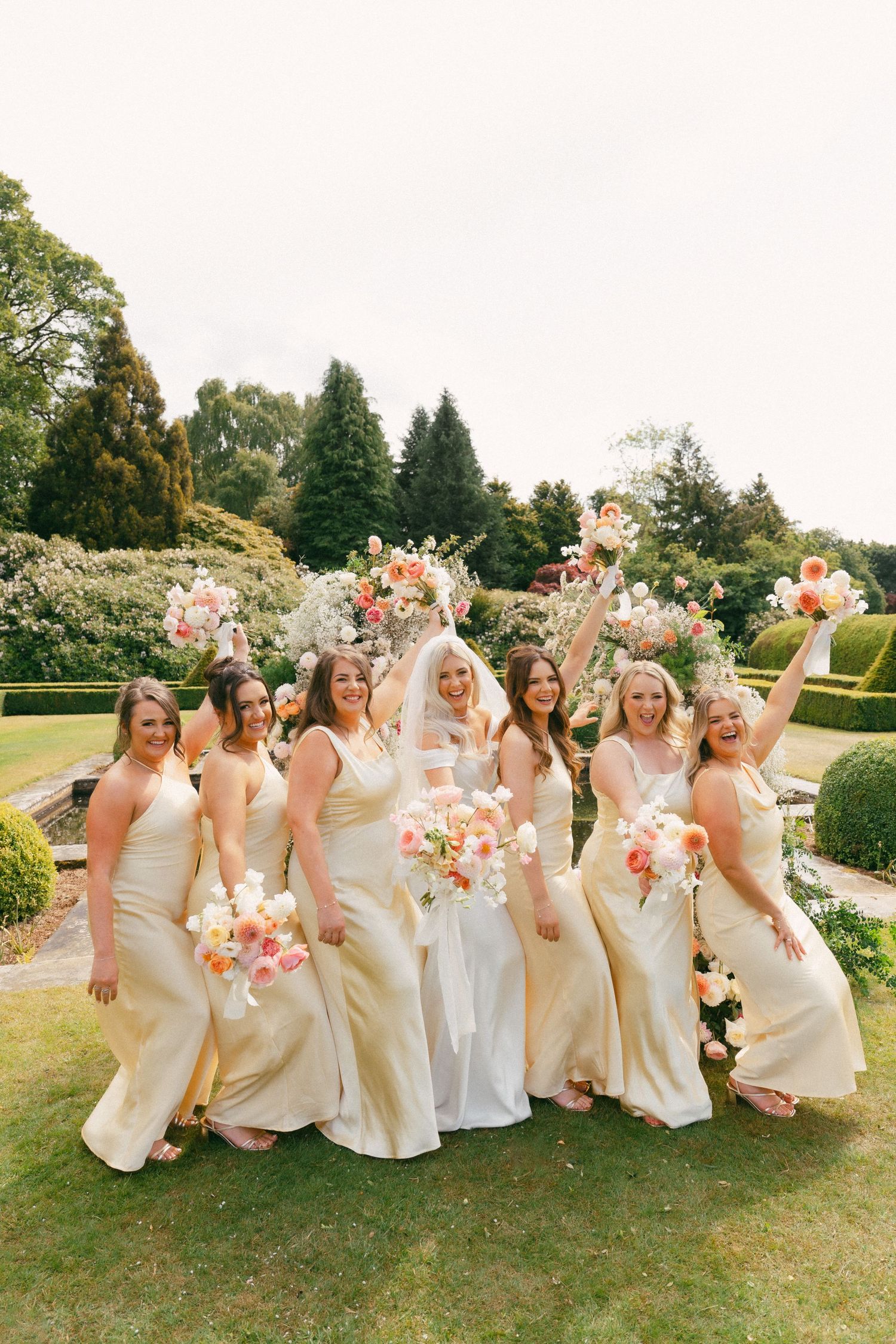 Wedding party in champagne-colored dresses celebrates joyfully on manicured garden lawn with pink floral bouquets.
