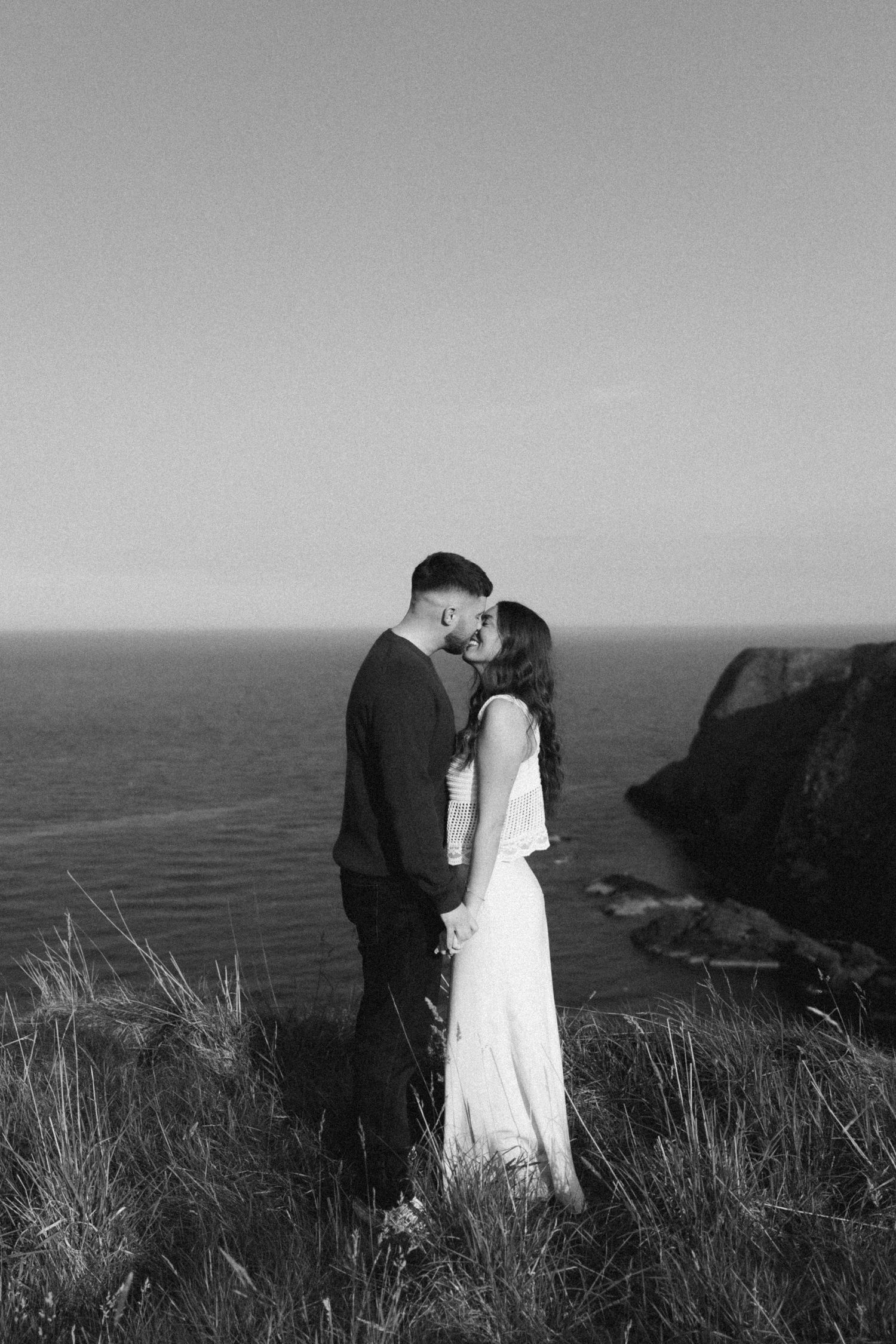 A romantic black and white coastal photo series showing a couple sharing intimate moments atop seaside cliffs.