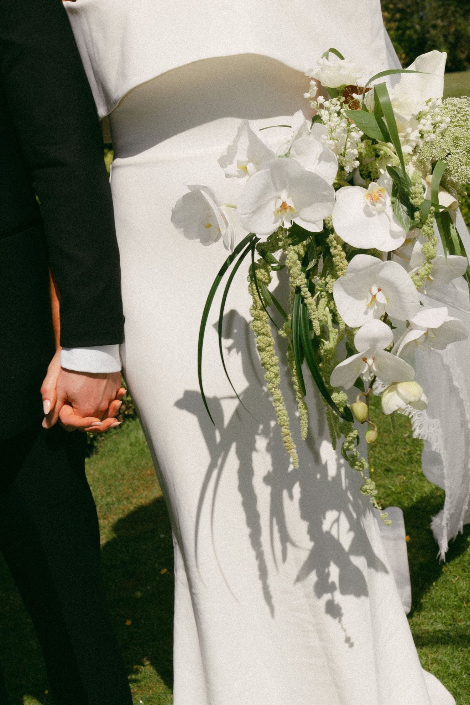 A bride and groom hold hands while a white orchid bouquet casts dramatic shadows on her elegant white wedding dress.