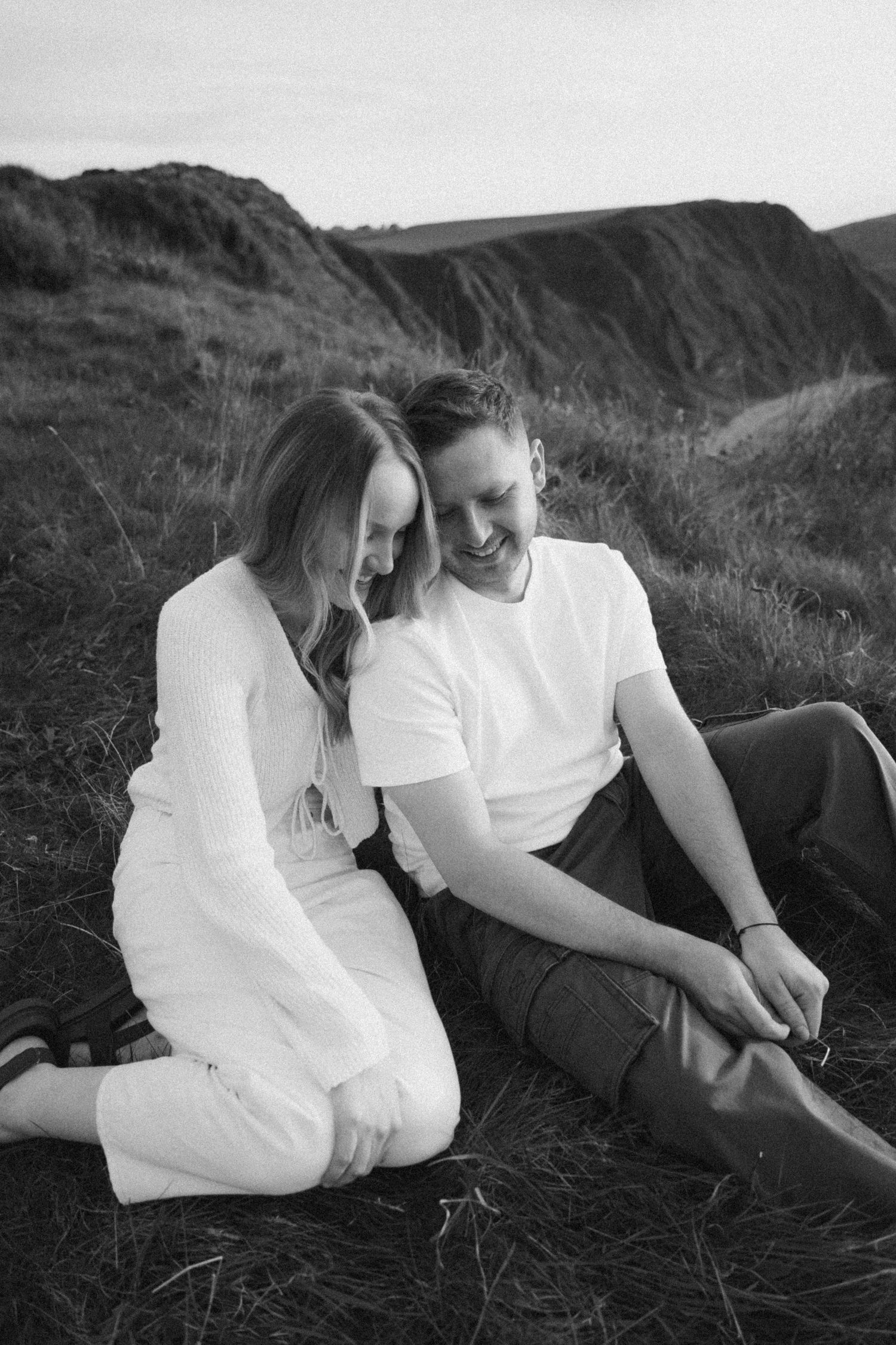 An intimate black and white portrait of a couple sitting on a coastal hillside.