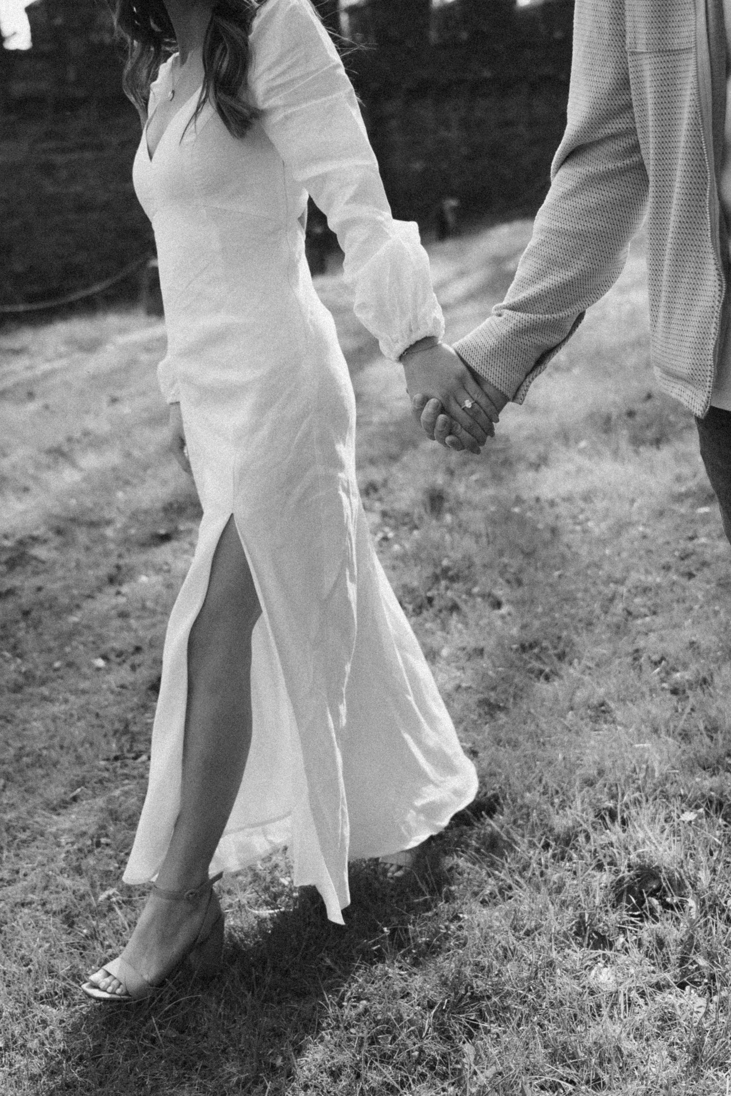 A couple holding hands while walking on grass, with the bride wearing a white dress with a side slit.