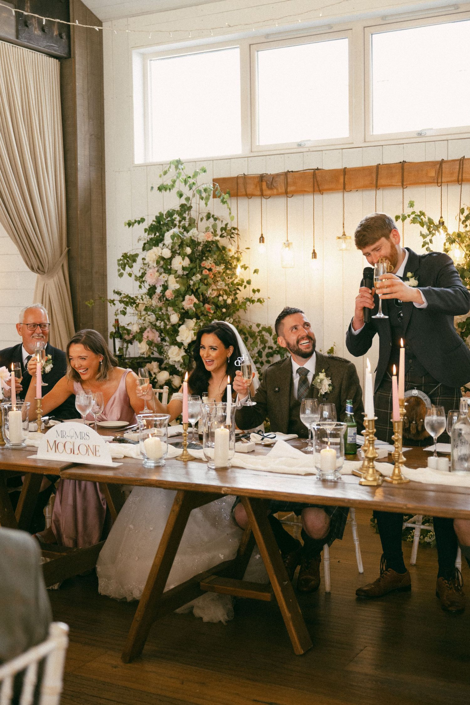 Wedding guests seated at a long wooden table enjoying dinner and drinks during an elegant reception celebration.