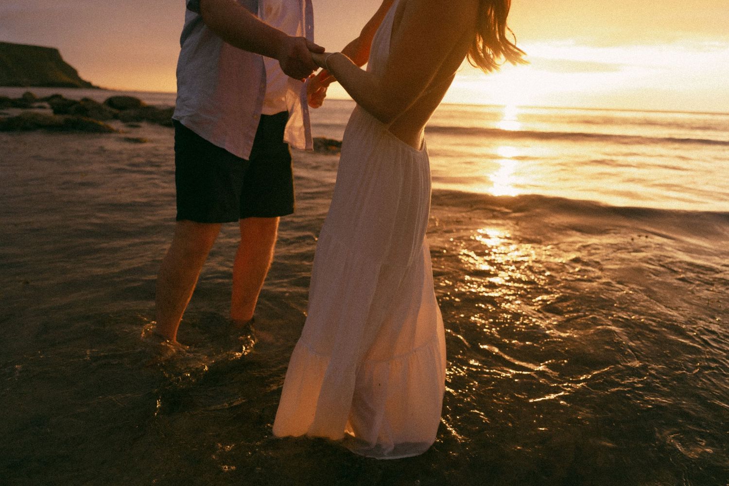 Two people standing in shallow ocean water at sunset with light reflecting off the waves.