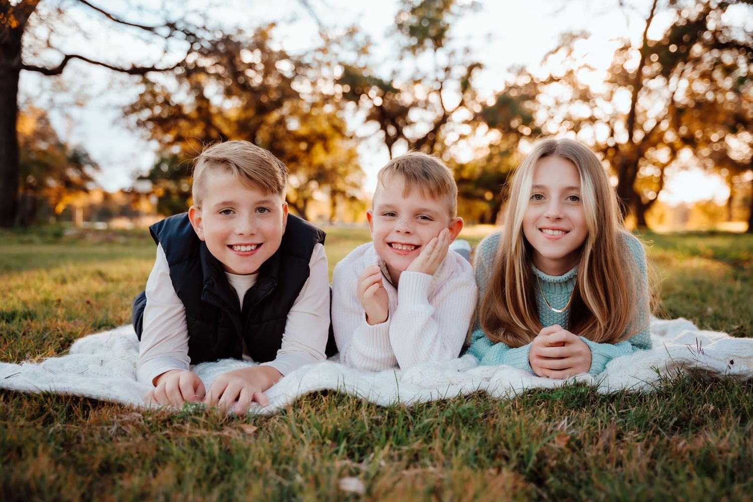 Three children lying on a blanket outdoors at sunset, smiling and enjoying a casual autumn photoshoot.