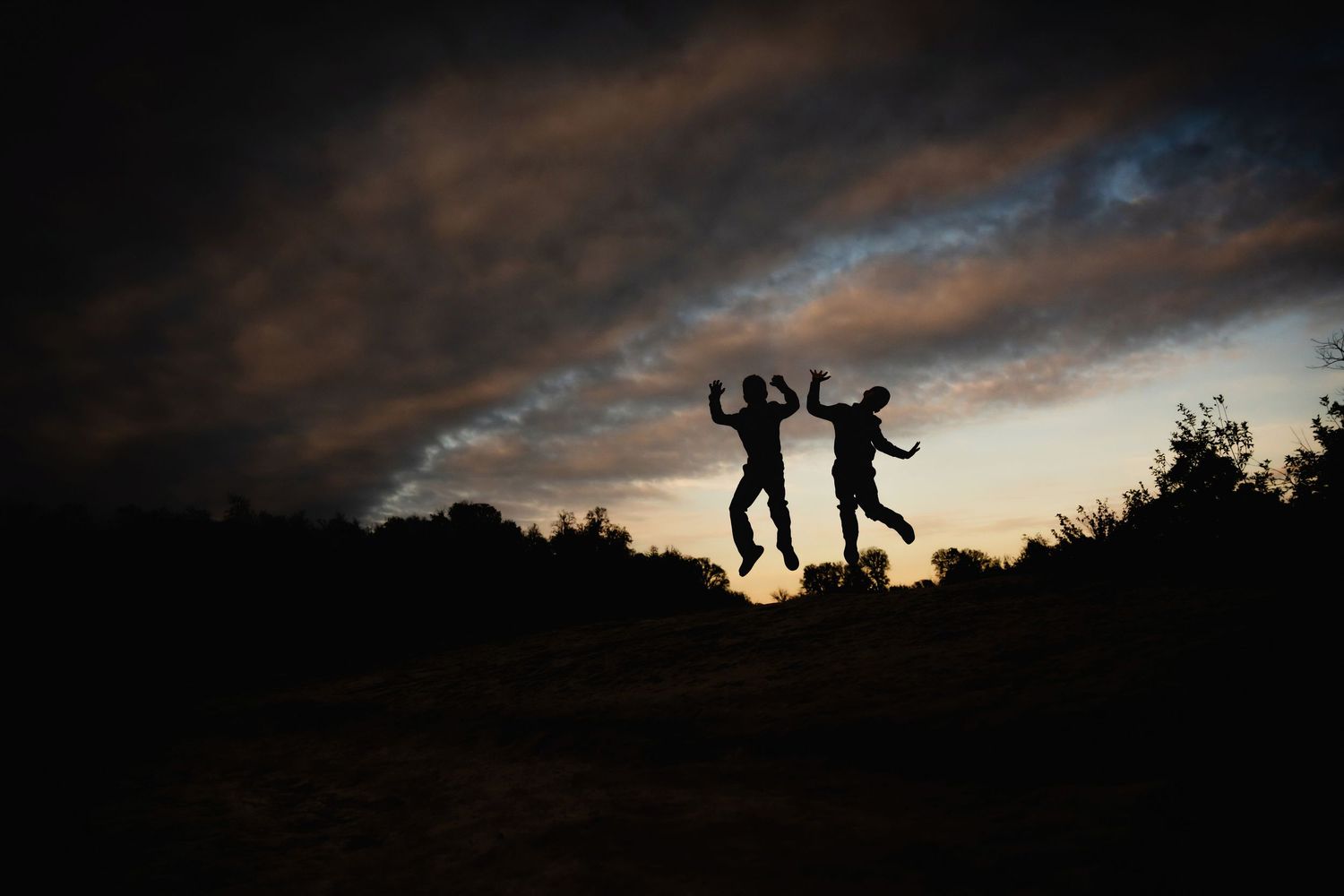 Silhouettes of two people dance joyfully against a dramatic sunset sky framed by dark trees in the background.