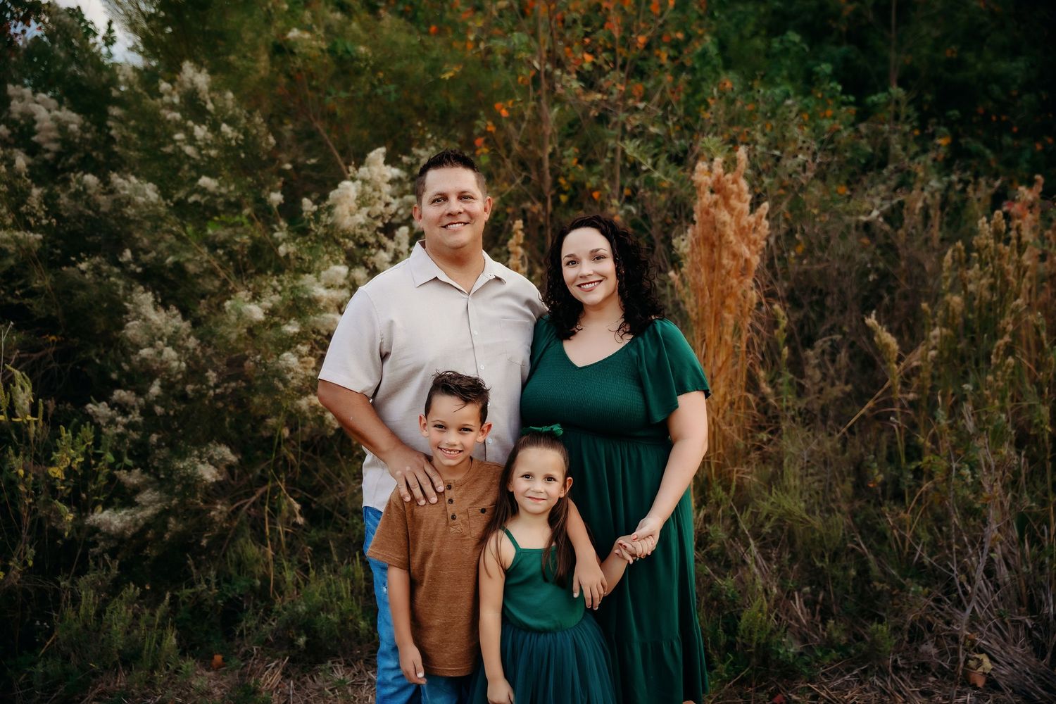 A family in coordinating green and beige outfits poses together outdoors against a natural backdrop of trees and foliage.