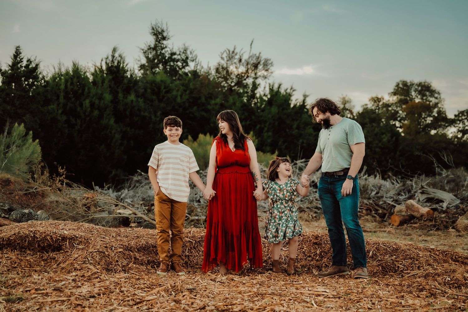 Three people stand together on a hilltop against evergreen trees during golden hour sunset.