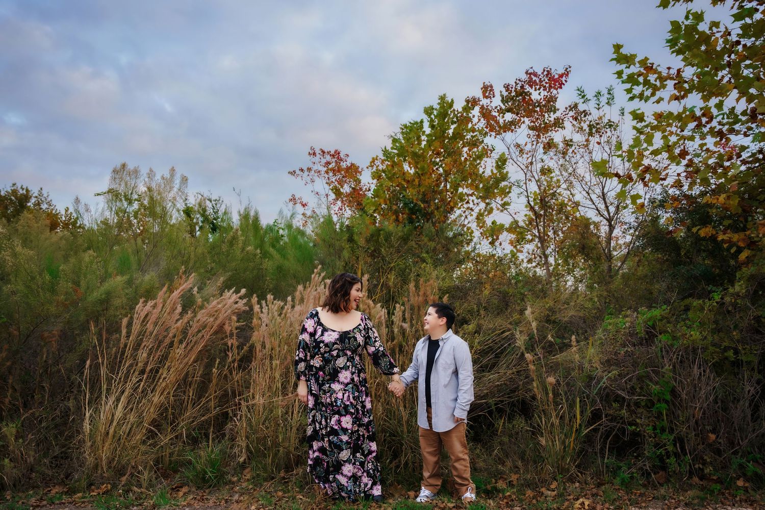 A couple stands together outdoors during sunset among autumn trees and tall grass.