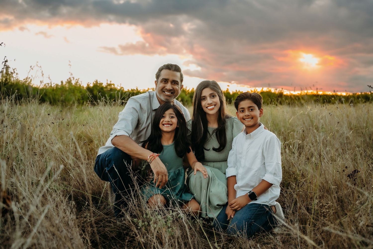 A group of four people sit together in a tall grass field during sunset with warm orange clouds in the sky.