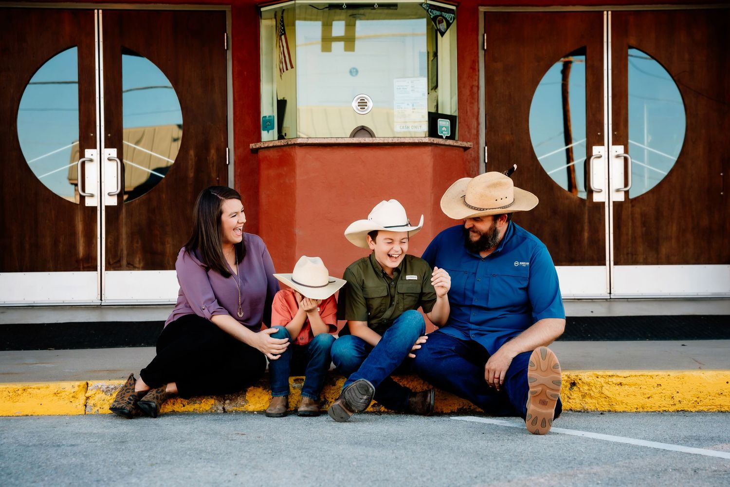 A family group sits together on the ground in front of a vintage storefront with circular windows and wooden doors.