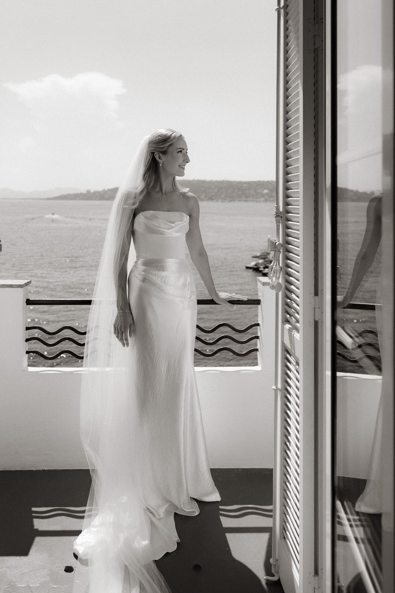 Black and white artistic photos of a bride in a white dress looking through shuttered windows at the ocean view.