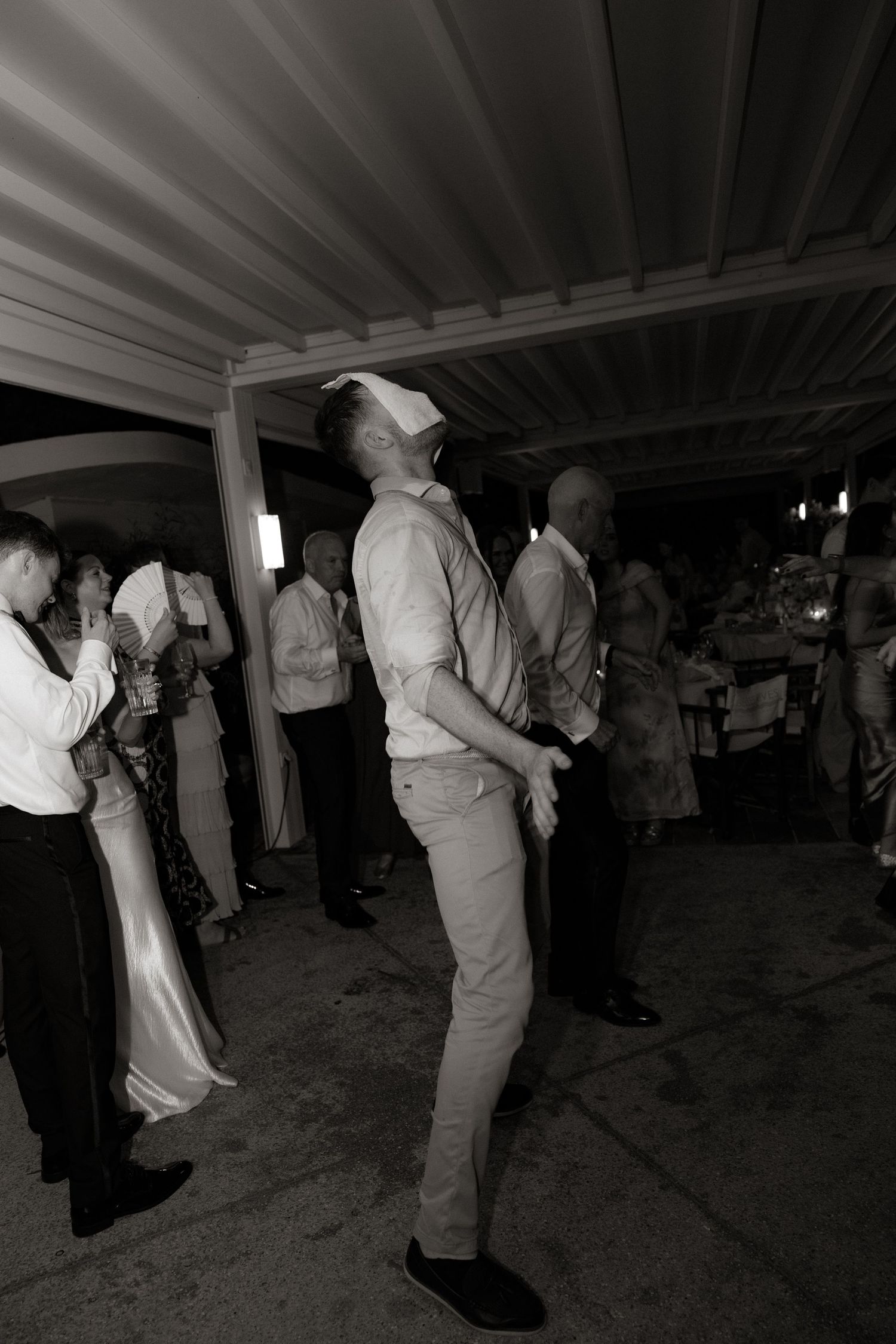 Black and white photo of people dancing and socializing at an indoor event with exposed ceiling beams.