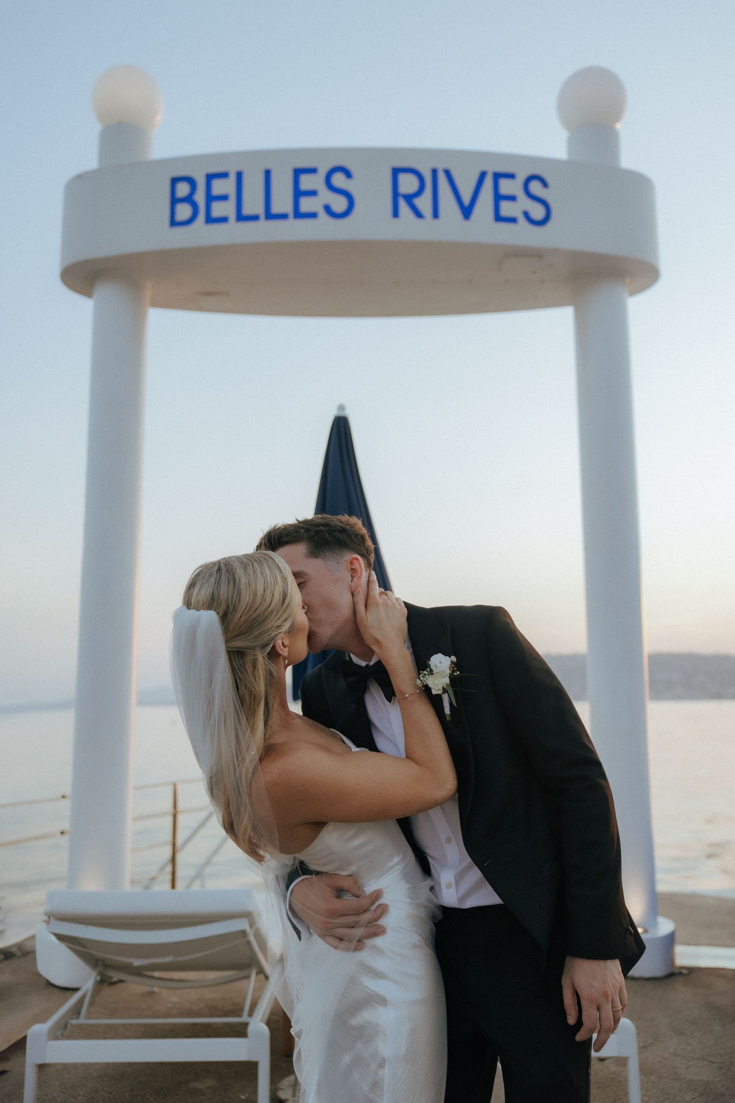 A wedding couple shares a romantic moment under a 'Belles Rives' archway at sunset.
