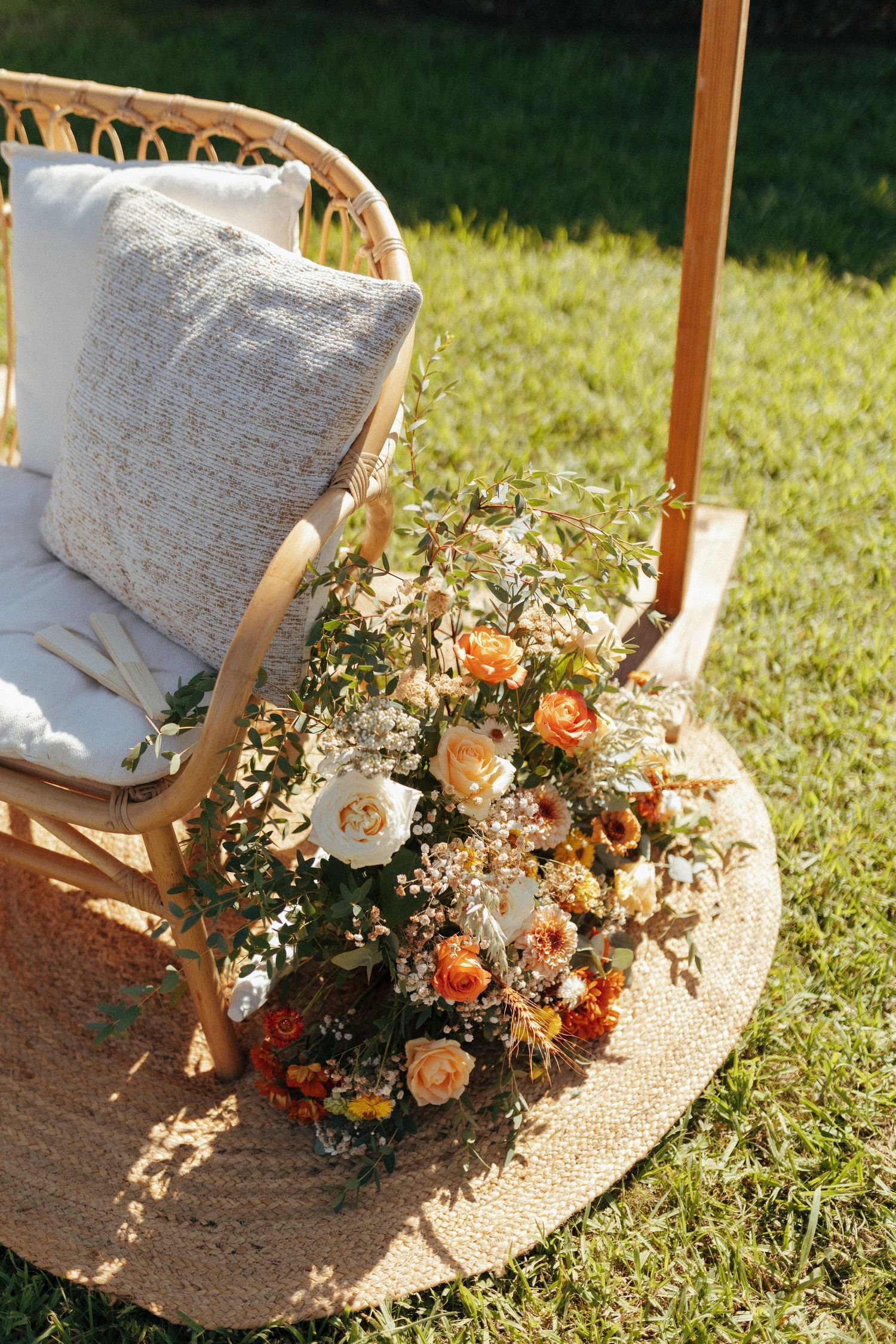 Outdoor wedding decor with rattan chair and colorful floral arrangement on jute rug in sunny garden setting.