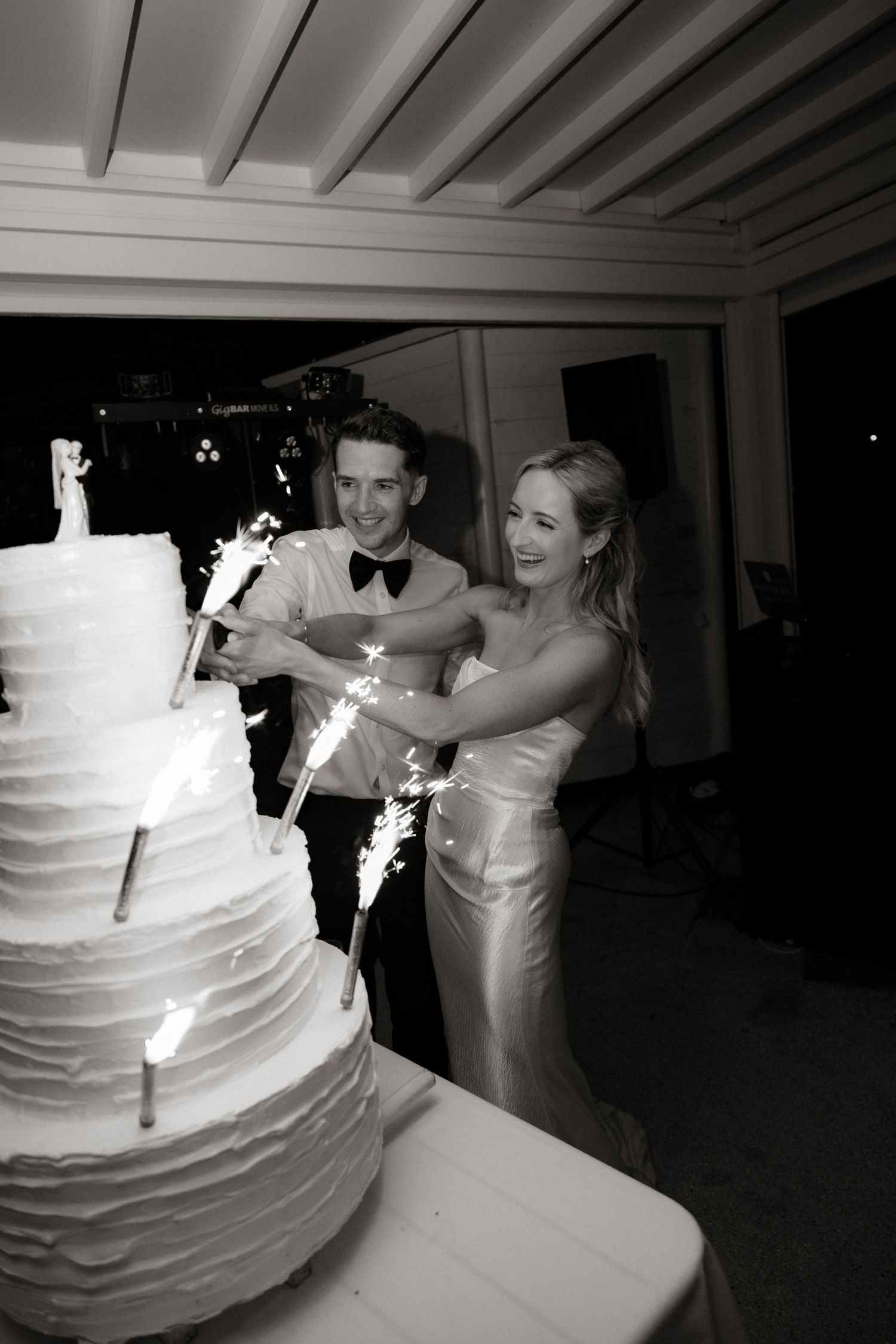 A newlywed couple cuts into their elegant white tiered wedding cake with sparklers in a black and white photo.