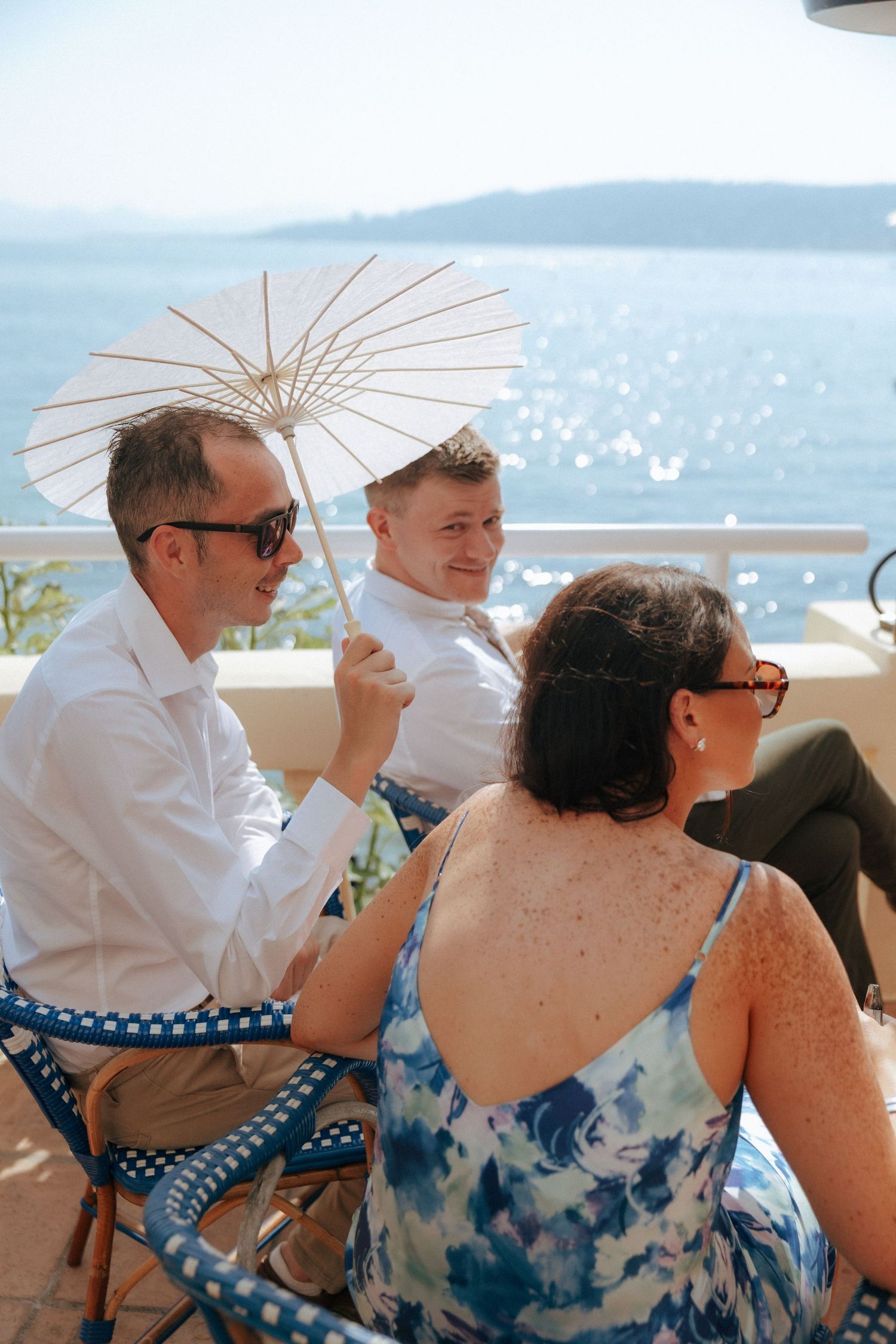 Guests enjoy a sunny outdoor wedding reception with a parasol and ocean views in the background.