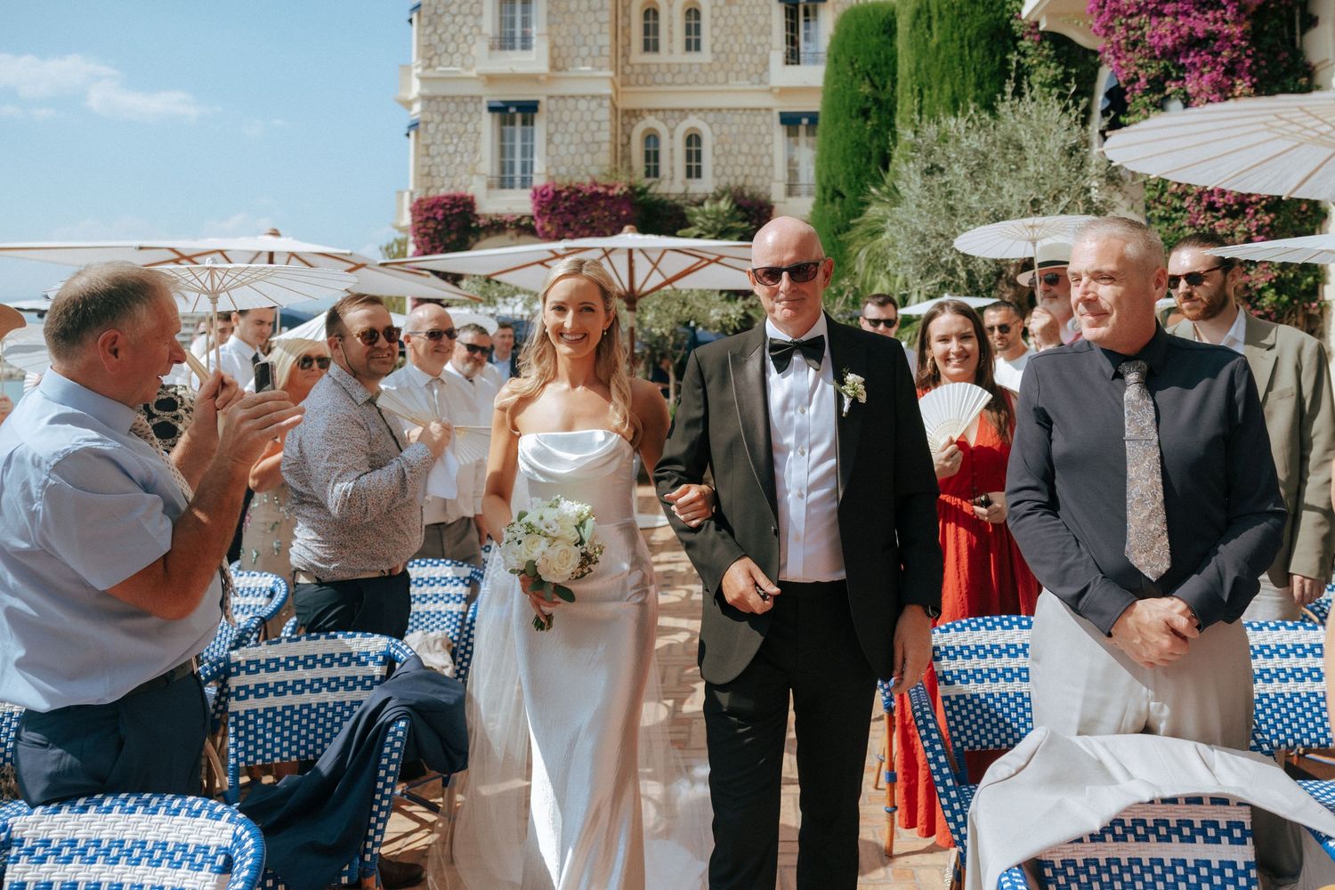 A wedding party walks through an outdoor Mediterranean restaurant terrace with blue and white chairs along the coast.