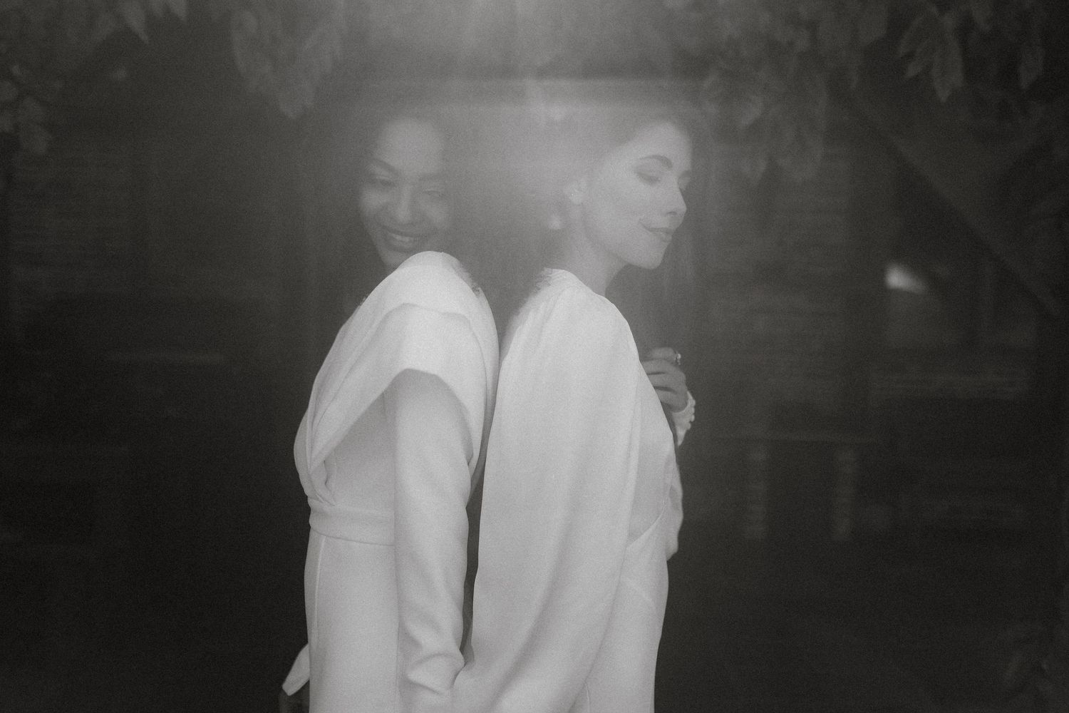 Two people in white shirts stand back-to-back in a moody black and white photograph with soft lighting effects.