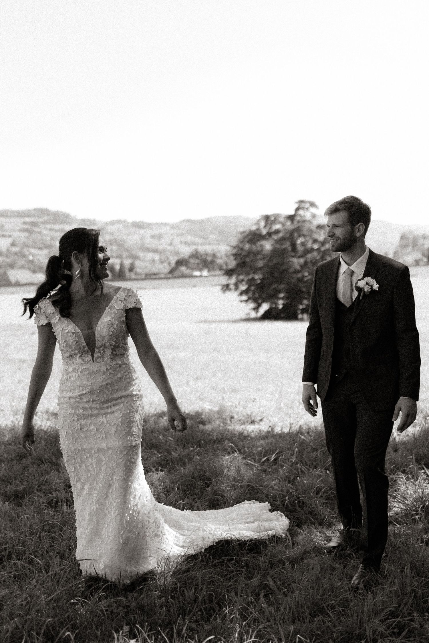 Black and white photo of a couple in wedding attire walking together in a scenic outdoor field.