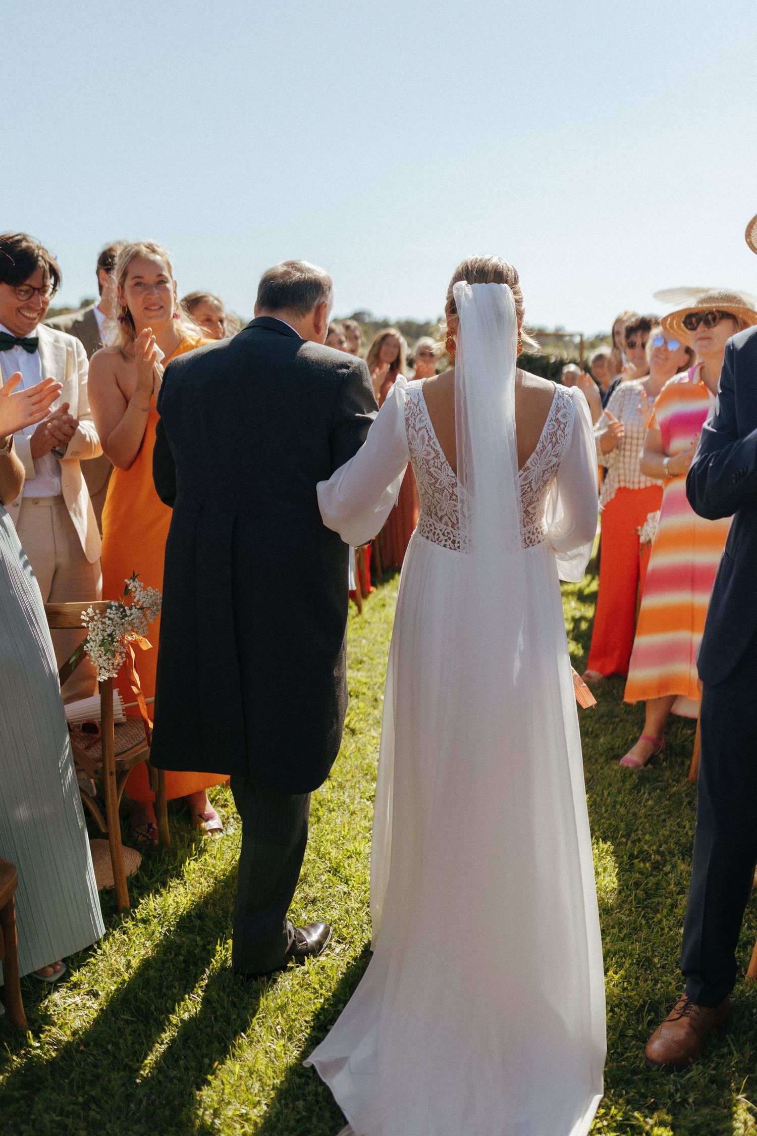 Guests applaud during an outdoor wedding ceremony as a bride in a delicate lace gown stands at the altar.