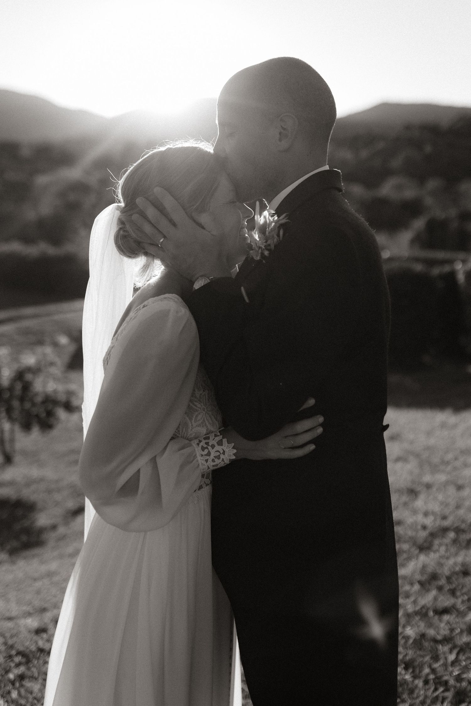 A romantic black and white wedding photo capturing a tender kiss between a couple in a sun-drenched outdoor setting.