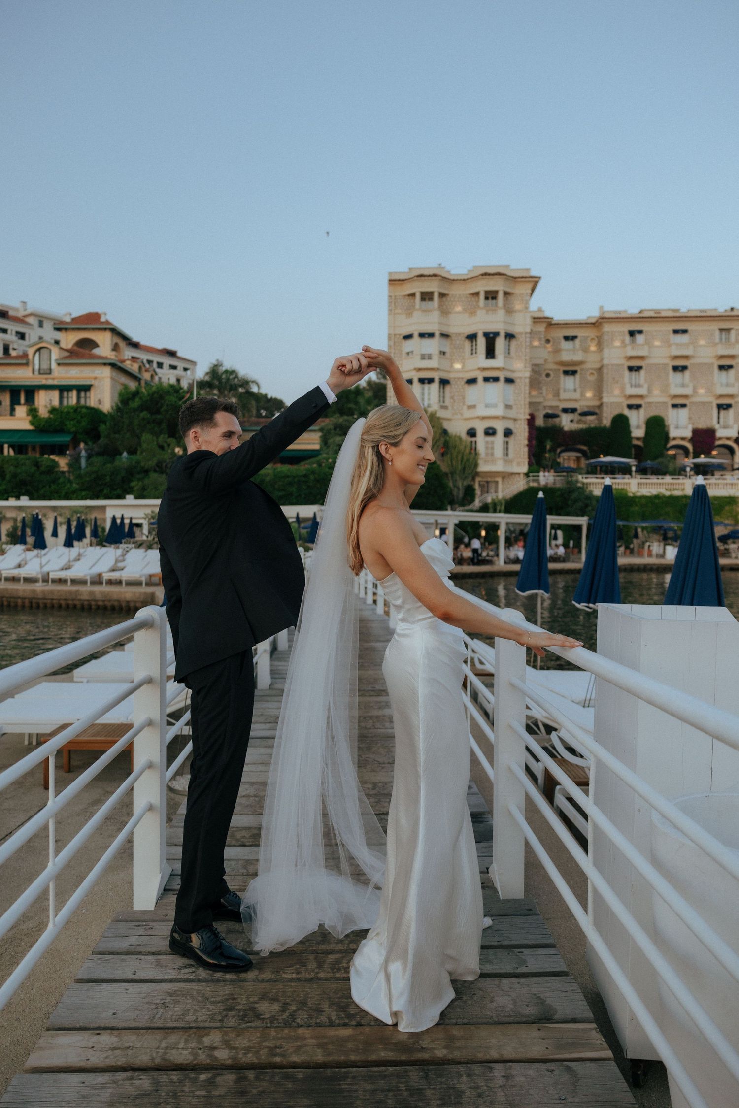 A wedding photoshoot on a wooden pier with coastal buildings visible in the distance.
